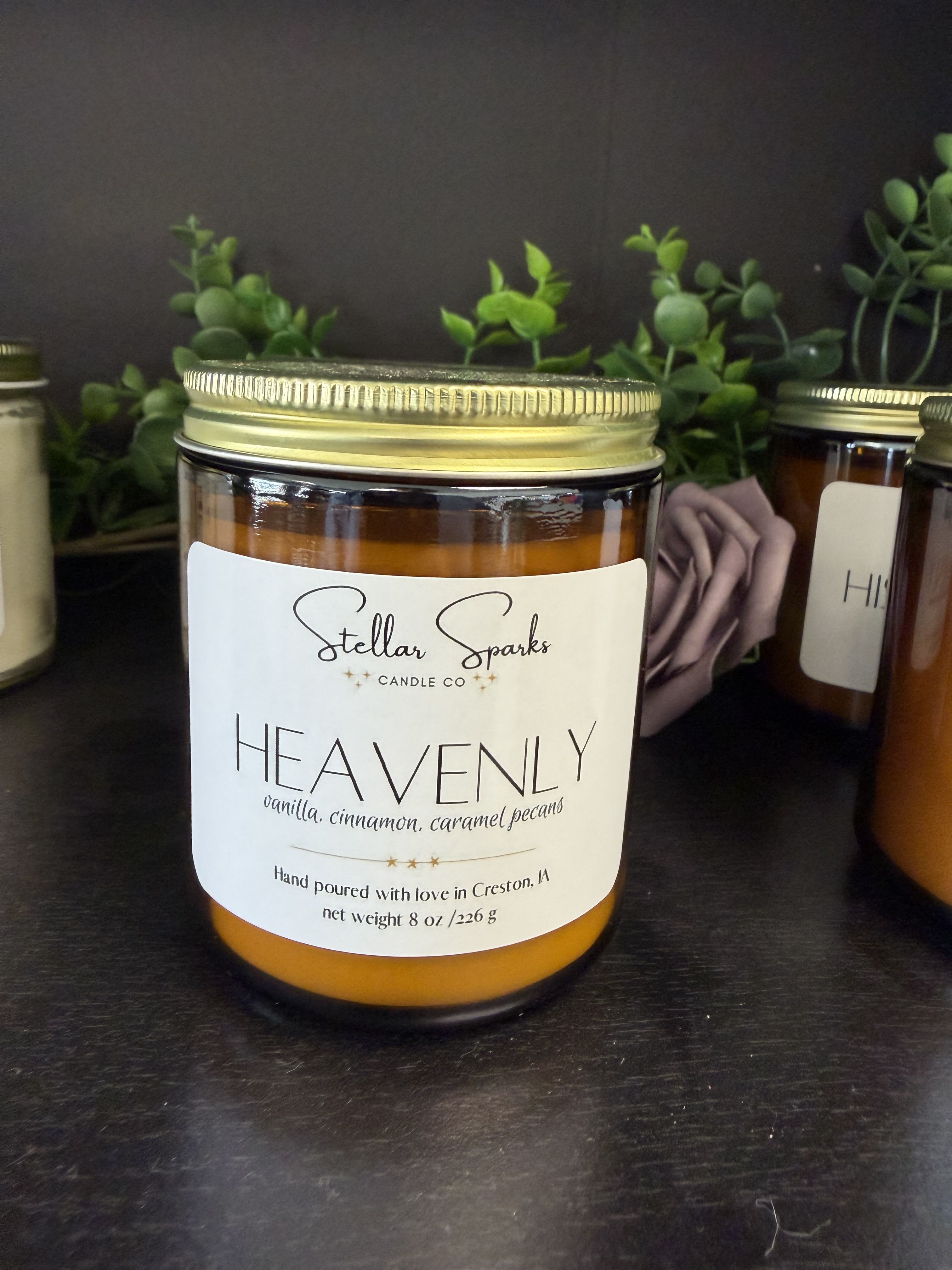 Heavenly 8oz Candle - A warm, cozy gift that reads like a comforting hug: this 8oz jar candle is perfect for seasonal decor, housewarming, or a thoughtful birthday present. The candle arrangement and presentation make it an ideal gift for someone who loves hygge moments or a touch of autumn warmth; the product type and occasion-gift-worthy candle for housewarming or seasonal celebrations-are clear from the first light.  The amber glass jar with a gold metal lid and a clean white label that reads "Heavenly" gives a classic, homey look that complements orange, white, black, and green accents in room decor. The scent profile-vanilla, cinnamon, and caramel pecans-suggests a sweet, spicy aroma that fills a room without overpowering it; faux greenery behind the jar in the display hints at how well it pairs with natural or seasonal styling. The printed label and sturdy jar make it feel polished enough for giving straight away.  Give this candle to someone who deserves a moment of calm-she'll notice the warm fragrance and the thoughtful presentation right away. The 8oz size is easy to place on a bedside table or mantel and comes topped with a metal lid to keep it fresh between uses, making it a simple, ready-to-give choice for holidays, thank-yous, or cozying up a new home.