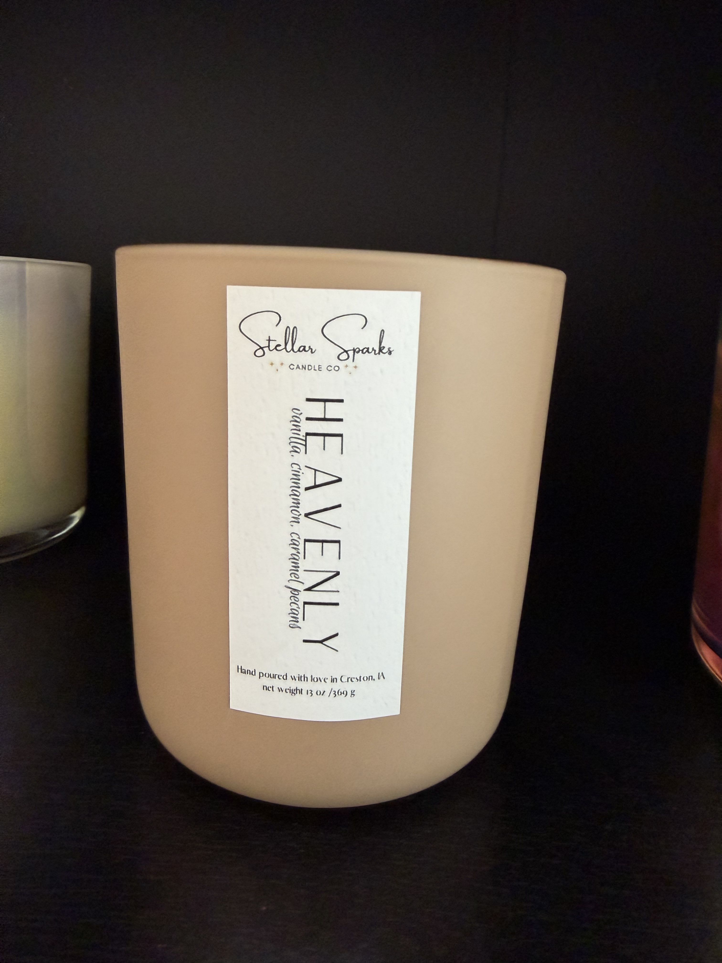 Heavenly 13oz Candle - A warm, comforting gift that reads like a hug in a jar-this 13oz home fragrance candle is ideal as a thoughtful housewarming or thank-you gift. The candle arrangement and packaging are intentionally understated, making it a suitable choice for birthday, hostess, or holiday gifting when you want the person you're thinking of to feel cozy and cared for.  The candle comes in a rounded beige ceramic-style jar with a tall white front label from Stellar Sparks Candle Co. The scent profile-vanilla, cinnamon, caramel, and pecans-creates a soft, gourmand aroma that fills a room without overpowering it. The neutral white, black, and beige palette and simple product label mean it fits easily into a range of décor styles, from minimalist to rustic.  Perfect for the recipient who prefers subtle luxury over flash, this candle is the kind of present that makes someone feel truly seen and comforted. The 13oz size offers several evenings of burn time; trim the wick between uses and place on a heat-safe surface away from drafts for best results.