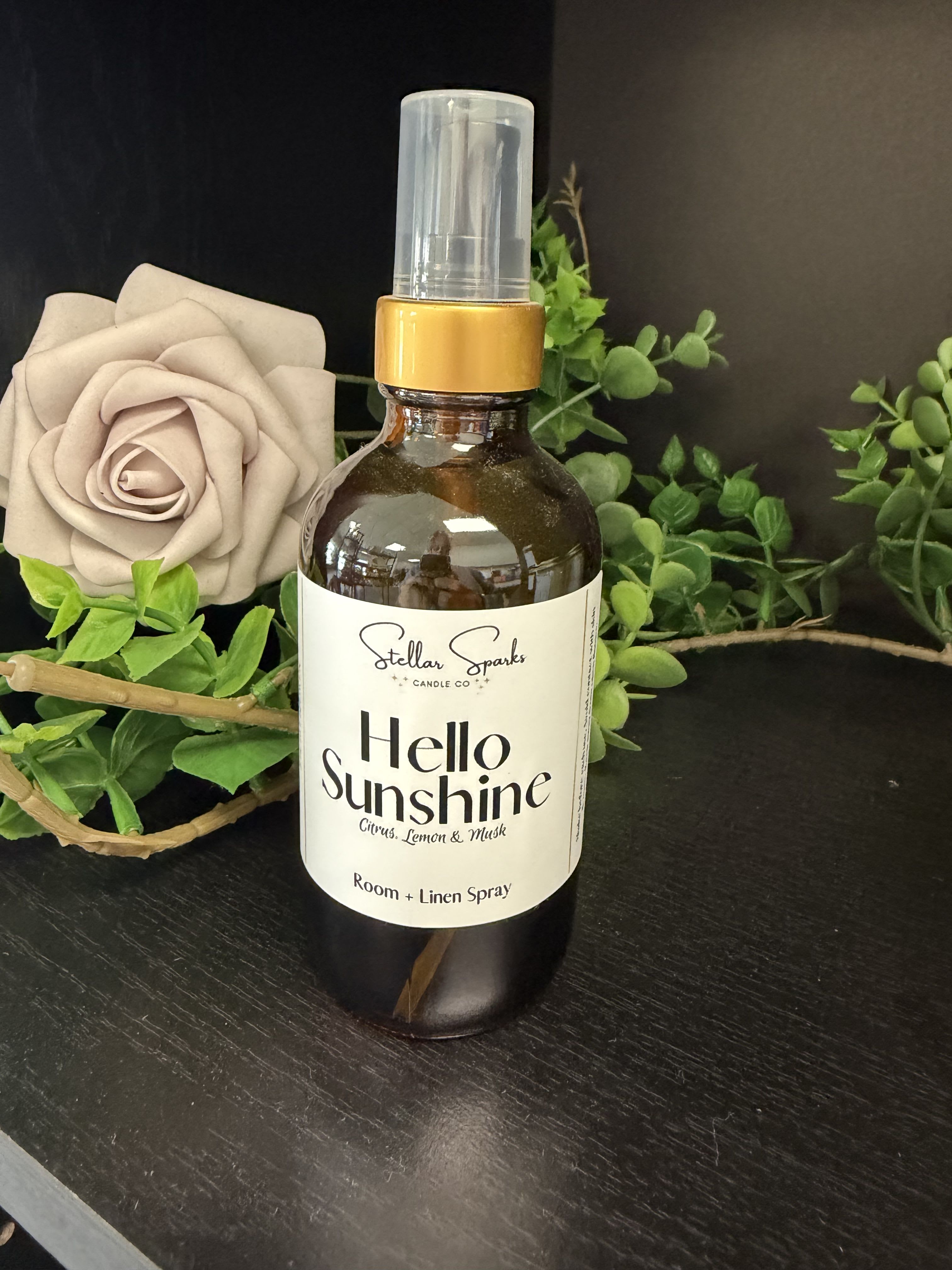Hello Sunshine Room Spray - "She'll notice the welcome scent first." This room and linen spray is a cheerful home fragrance gift-perfect as a housewarming present, birthday add-on, or a small way to say you're thinking of her. The amber spray bottle labeled "Hello Sunshine" reads bright and friendly on a mantel or bedside table, promising a fresh, uplifting aroma that makes a room feel cleaner and more inviting.  Styled with a large beige rose and green sprigs, the arrangement highlights soft natural tones of beige, white and green against the bottle's black-accented cap and label. The product includes the amber spray bottle, plastic cap, and product label shown; the rose in the image is decorative styling to convey the scent's floral character. Compact and easy to use, the spray is ready to refresh linens, curtains, or the air in small rooms-an uncomplicated, joyful way to make the person you're gifting feel noticed and at home.