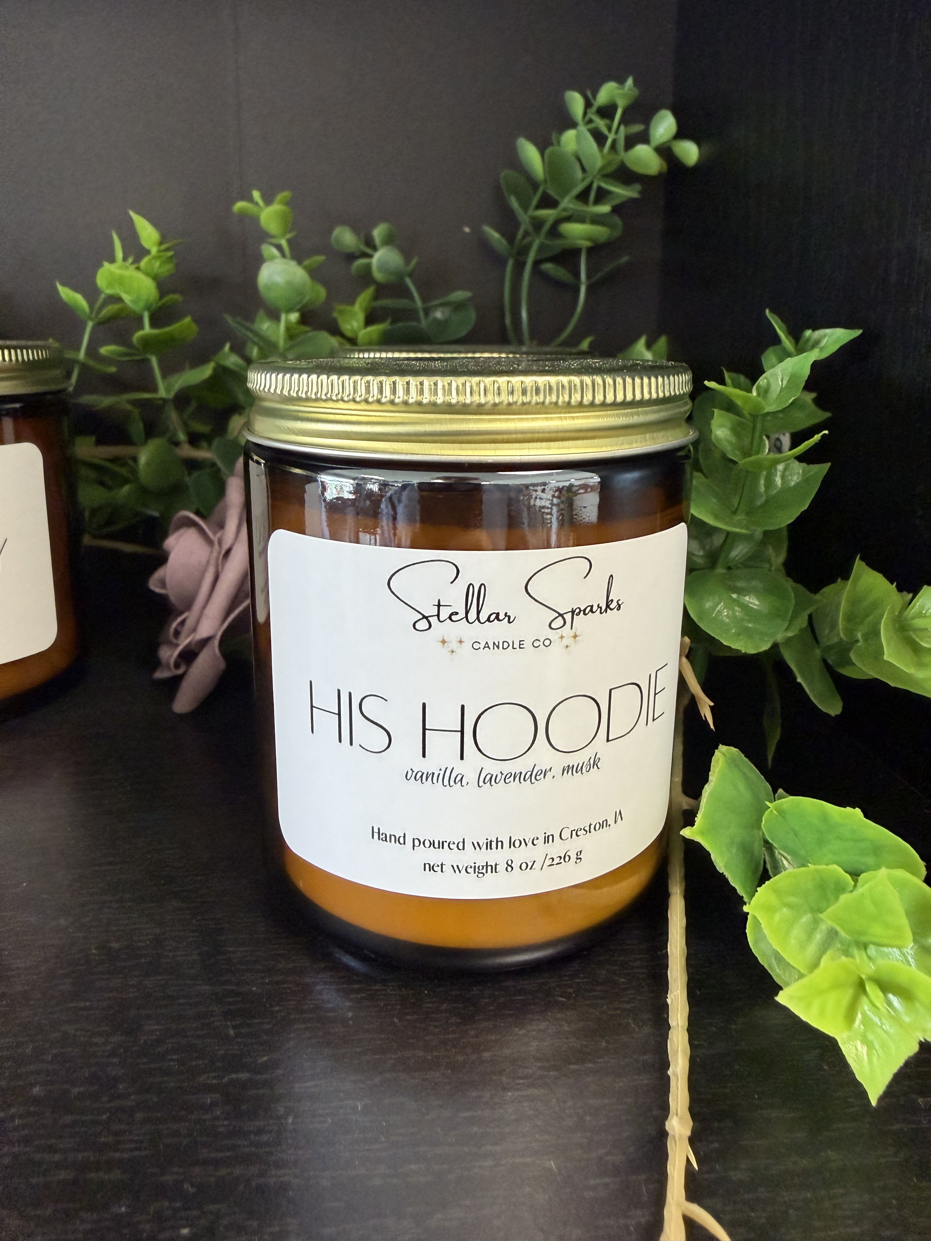 His Hoodie 8oz Candle - The warm, familiar scent of "His Hoodie" greets the room with cozy vanilla, soothing lavender, and soft musk-a gift-ready home fragrance that's perfect as a masculine-leaning birthday or "thinking of you" present. As a housewarming or anniversary add-on, this 8oz scented candle offers the comforting impression of a loved one's sweater wrapped around the person you're gifting it to, making them feel cared for and remembered.  Housed in an amber glass jar with a gold metal lid and a large white label, the candle arrives styled with decorative faux greenery for a rustic, lived-in look that fits modern or farmhouse decor. The clean, understated packaging keeps the focus on scent while making it easy to pair with floral gifts or give on its own. Burn time for an 8oz jar is ideal for several evenings of fragrance; follow standard candle-safety care-trim the wick, keep away from drafts, and never leave burning unattended.