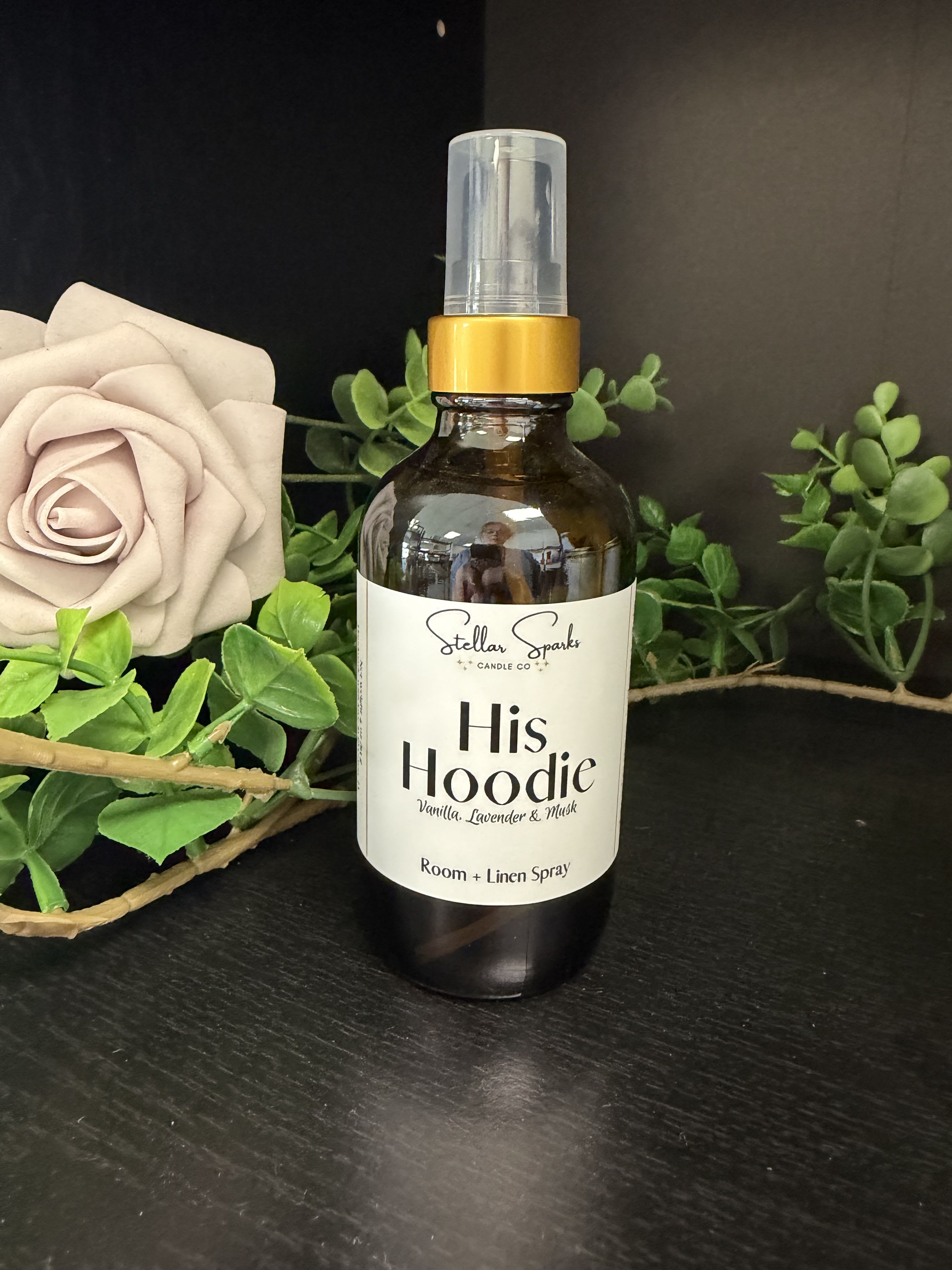 His Hoodie Room Spray - Cream rose petals and bright green foliage soften a rich amber bottle in this room-and-linen spray-an intimate home fragrance gift perfect for anniversaries, birthday treats, or "just because" moments. As a scent-forward present for him or her, the spray pairs the quiet romance of roses with the lived-in comfort of a favorite hoodie or pillow, making the person you're thinking of feel noticed and cozy.  The arrangement features a single large cream rose and vivid green leaves artfully placed beside an amber glass spray bottle with a white label and gold collar, finished with a plastic spray nozzle. The dark background highlights the bottle's warm tones and the rose's pale texture, creating a calm, slightly romantic aesthetic suitable for bedside tables, linen closets, or entryway styling.  Gift this spray when you want to evoke warmth and familiarity-perfect for couples, close friends, or anyone who appreciates subtle floral notes in their home. Bottle size and scent concentration are shown on the label; spritz lightly on fabrics and in rooms, avoiding direct contact with delicate materials or finished wood surfaces.