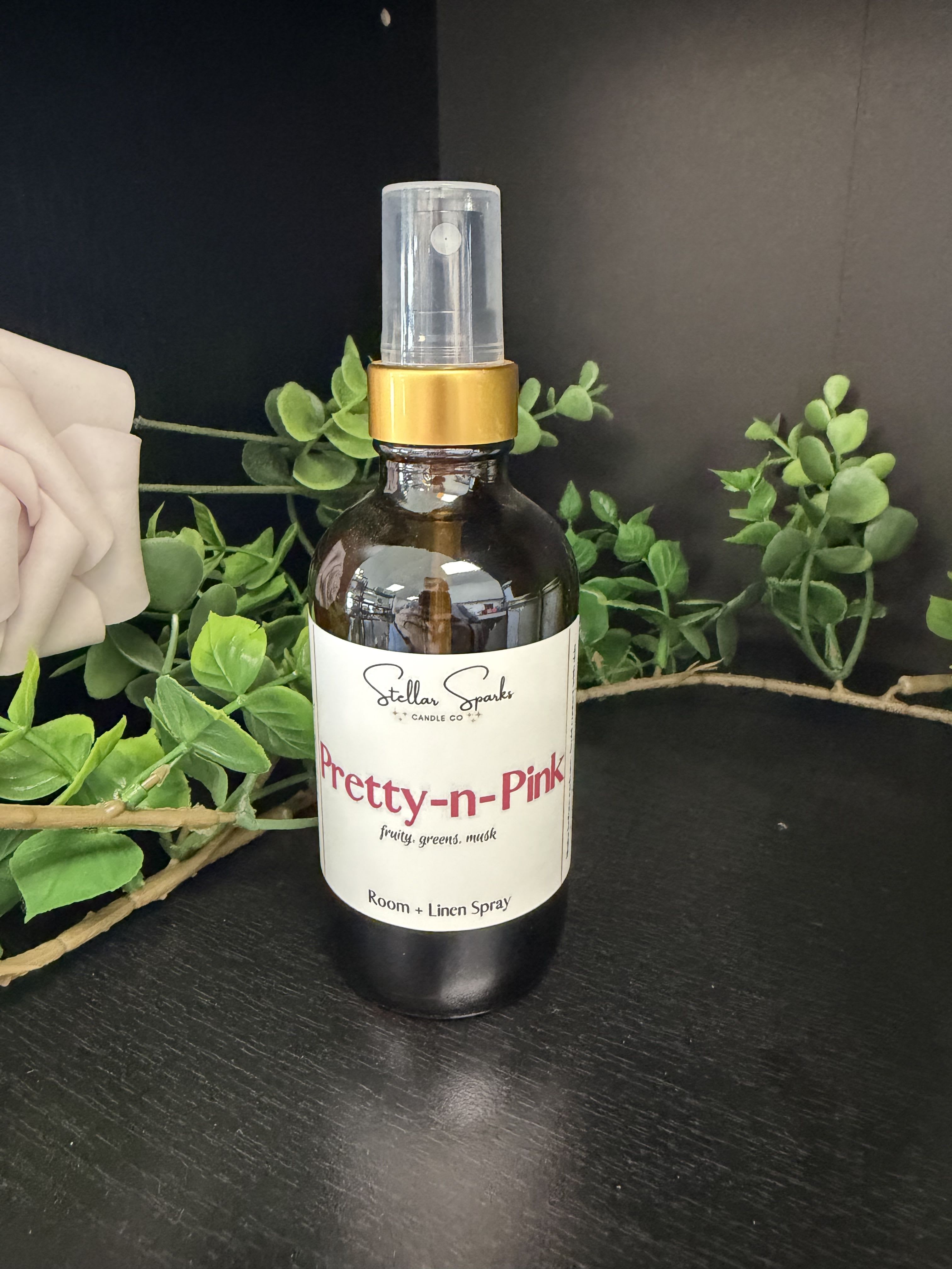Pretty-N-Pink Room Spray - A delicate, feminine room and linen spray that reads like a bouquet in a bottle - the Pretty-N-Pink room spray is perfect as a giftable home accent or a finishing touch for a birthday, Mother's Day, or a just-because surprise. The scent leans fruity with green and musky notes, designed to make the person you're thinking of feel refreshed and gently celebrated the moment they walk into the room.  The spray comes in a dark amber bottle with a gold collar, clear plastic cap, and a clean product label that fits boutique styling on any dresser or entry table. Styled here against pale pink florals and soft greenery, the colors-pink, white, green and black-signal a softly romantic, modern aesthetic. Use it on linens, in bathrooms, or as a quick air refresh; the included plastic spray cap gives an even mist for light, long-lasting fragrance.