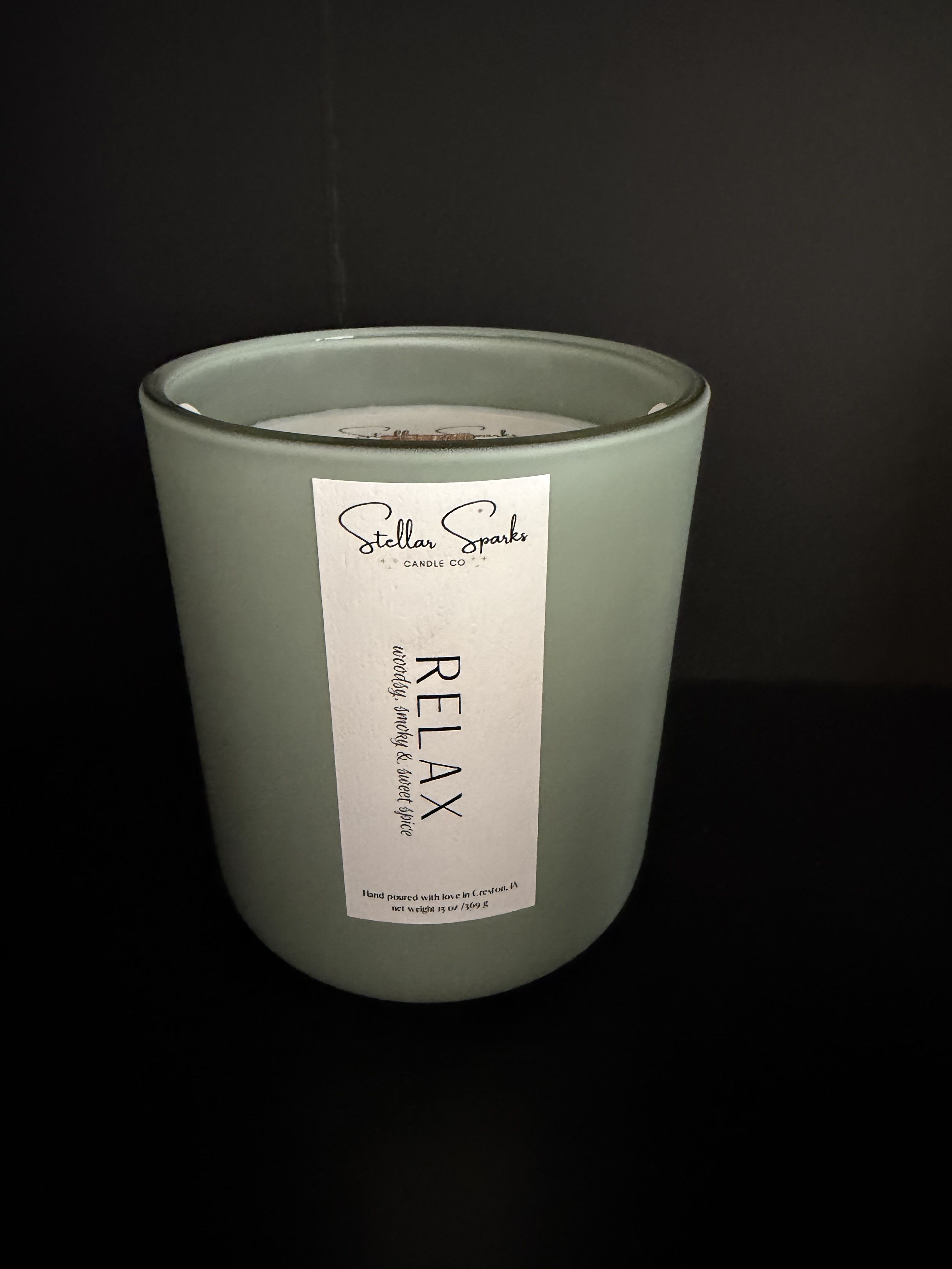 Relax 13oz Candle - Muted sage green glass and a clean white label make this 13oz Relax candle a quietly elegant gift for anyone who needs a moment of calm. As a home fragrance or small birthday, hostess, or "thinking of you" present, the arrangement-like simplicity of this candle feels intentional-she'll notice the soothing color and the understated "RELAX" label before the first warm, woody breath of scent settles the room.  The candle comes in a cylindrical glass jar in soft green with a white paper front label marked RELAX and a note of woody, smoky, sweet spice fragrance. The minimal black-and-white typography and matte glass give it a refined, modern look that suits living rooms, bathrooms, or bedside tables. At 13 ounces, it offers substantial burn time while remaining compact enough to tuck into a gift box or pair with a small bouquet.  Perfect for someone who appreciates calm, low-key design, this candle sends a thoughtful, restorative message-ideal for new homeowners, busy friends, or anyone needing a peaceful break. Place on a stable surface, trim the wick to 1/4" between uses, and enjoy the layered woody-smoky-spice scent as it fills the space.