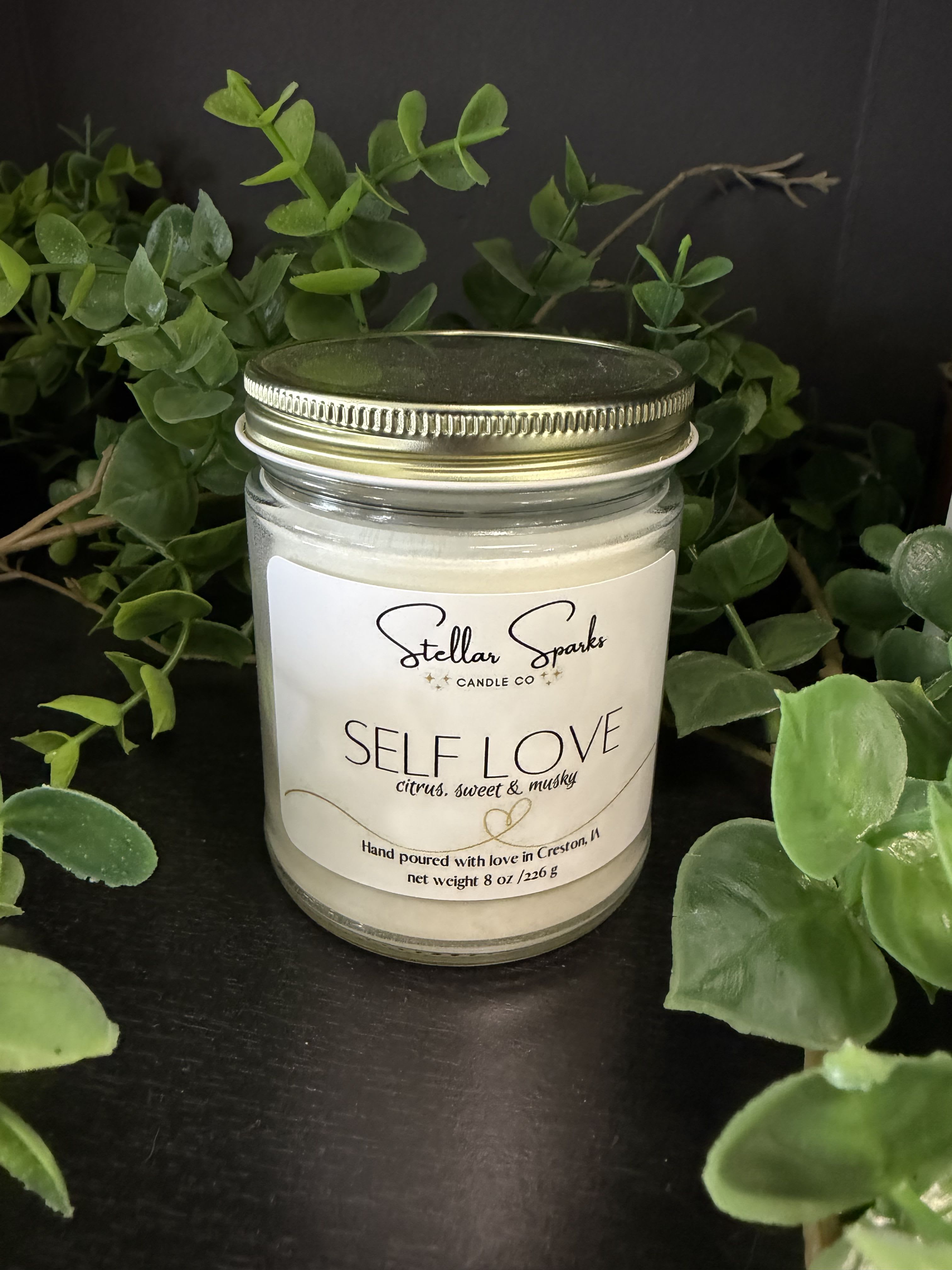 Self Love 8oz Candle - A calming, boutique-style glass jar candle that sets a gentle, self-care mood-perfect as a gift or a personal indulgence for moments of reflection. This Self Love 8oz candle is presented as a mindful gift item and works beautifully for occasions like birthdays, anniversaries, or simply to say "take care of yourself." The green foliage backdrop and clean, minimalist styling give it a fresh, restorative presence that helps the recipient feel seen and soothed.  The candle comes in a clear glass jar with a gold-toned metal lid and a crisp white label reading "Self Love." Cream-colored wax and a simple, handmade look make it feel thoughtful and refined; color accents are green, white, and black in the presentation. Compact at 8oz, it's easy to display on a bedside table or bathroom shelf and reads as both decorative and practical.  Give this as a small, meaningful way to encourage rest and self-appreciation-the kind of gift that says you want someone to slow down. Ideal for sending to a friend, partner, or anyone in need of a quiet pick-me-up; order as a standalone treat or pair with a bouquet for a fuller care package.