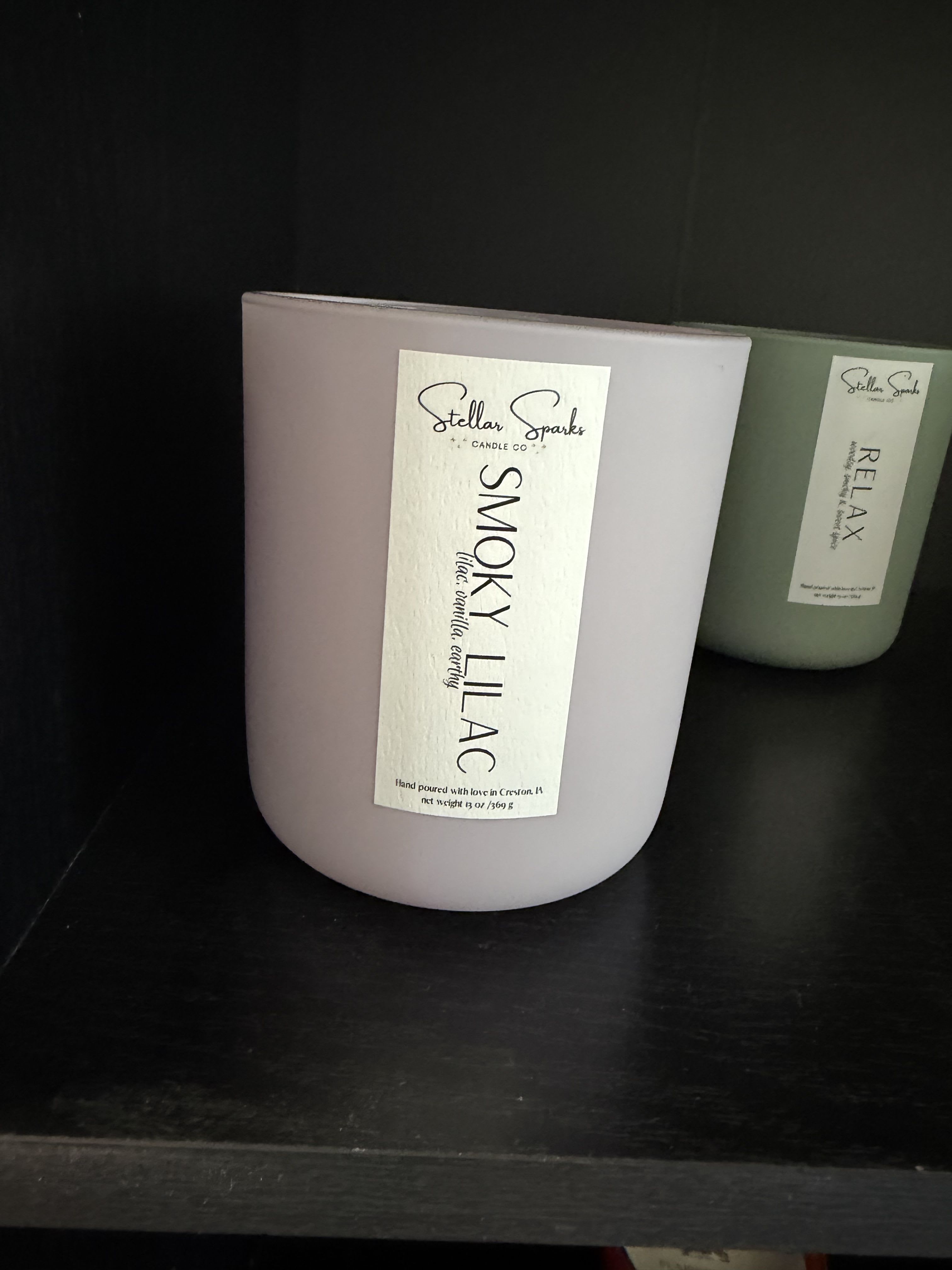Smoky Lilac 13oz Candle - Soft lavender and smoky white combine in a calming home-fragrance gift that reads like a floral moment in a jar-perfect as a thoughtful birthday token or a serene "thinking of you" present. This scented candle arrangement pairs the product type (scented candle) with the occasion context (gift for birthdays or quiet celebrations) right away so the person you're buying for feels noticed and soothed.  The 13oz Smoky Lilac candle comes in a matte ceramic jar with a clean printed label, the soft purple hue softened by touches of white, green, and black in the styling for a spa-like, contemporary look. The scent profile centers on lilac with a subtle smoky edge that evokes floral sweetness tempered by warmth; the simple packaging and muted tones make it an elegant addition to a bedside table, bathroom, or living room shelf.  Give this to someone who appreciates quiet luxury and a restorative atmosphere-the kind of gift that helps them slow down and breathe. The ceramic jar is reusable after the wax is gone; for best scent performance, trim the wick to 1/4" before each burn and allow the wax to form a full melt pool on the first burn.