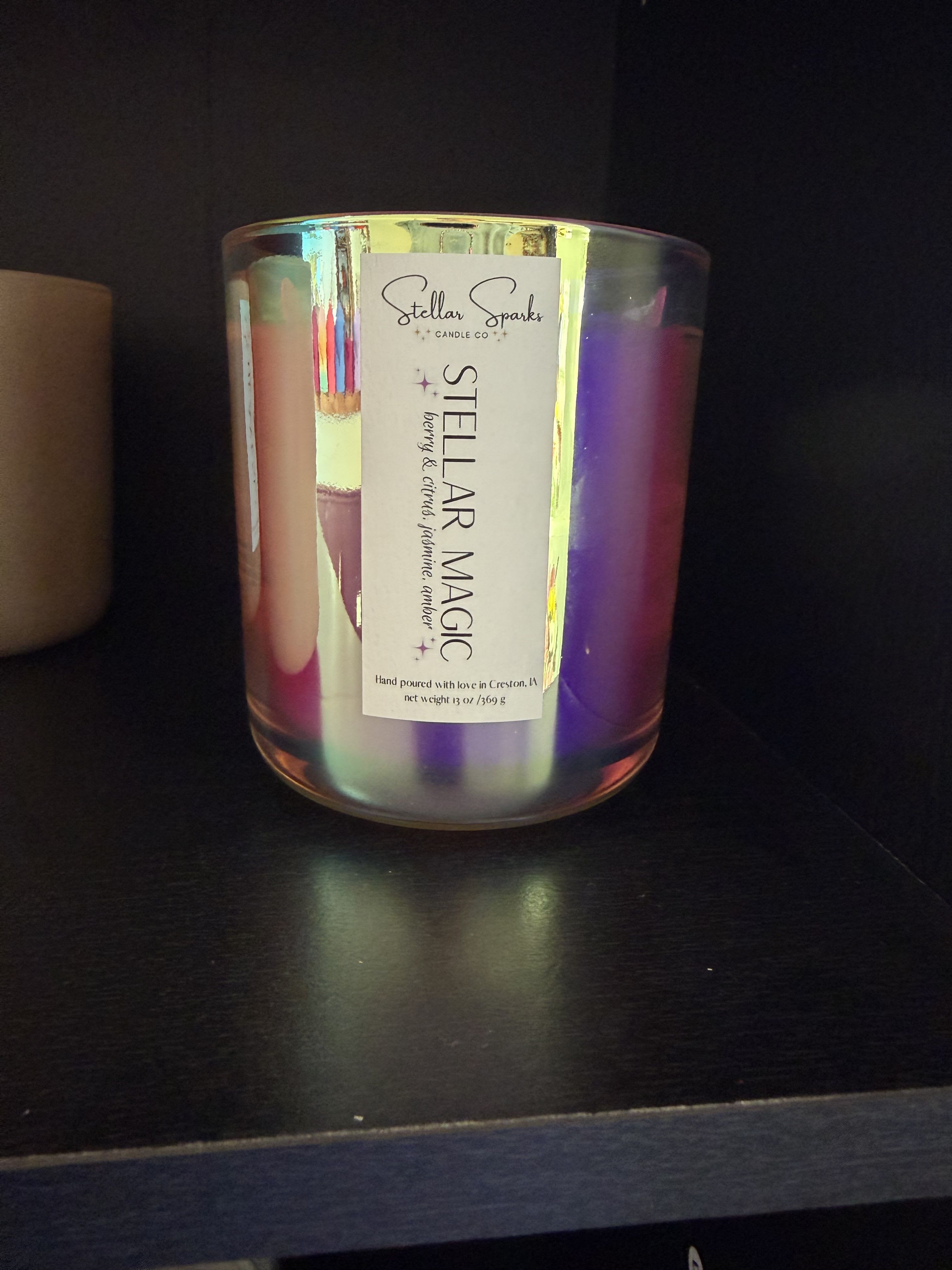 Stellar Magic 13oz Candle - Iridescent purple and pink glass catches the light like a little aurora-perfect as a gift for someone who loves cozy, playful home accents. This candle is a 13oz scented candle presented in a clear, reflective glass jar labeled Stellar Sparks Candle Co.; the product reads as a thoughtful housewarming or birthday gift that brings a warm, fragrant glow to any room.  The fragrance blends berry and citrus top notes for a bright, fruity scent that balances sweet and fresh. The jar's shifting purple, pink and pearlescent tones make it decorative even when unlit; a simple paper label names the scent and keeps the look modern and gift-ready. The candle is ideal for bedroom, living room, or as a centerpiece on a side table.  Give this as a cheerful way to say "thinking of you," celebrate a small milestone, or add a touch of sparkle to someone's home. Size: 13oz in a glass jar; ships ready to gift with its clean paper label-no extra styling needed.