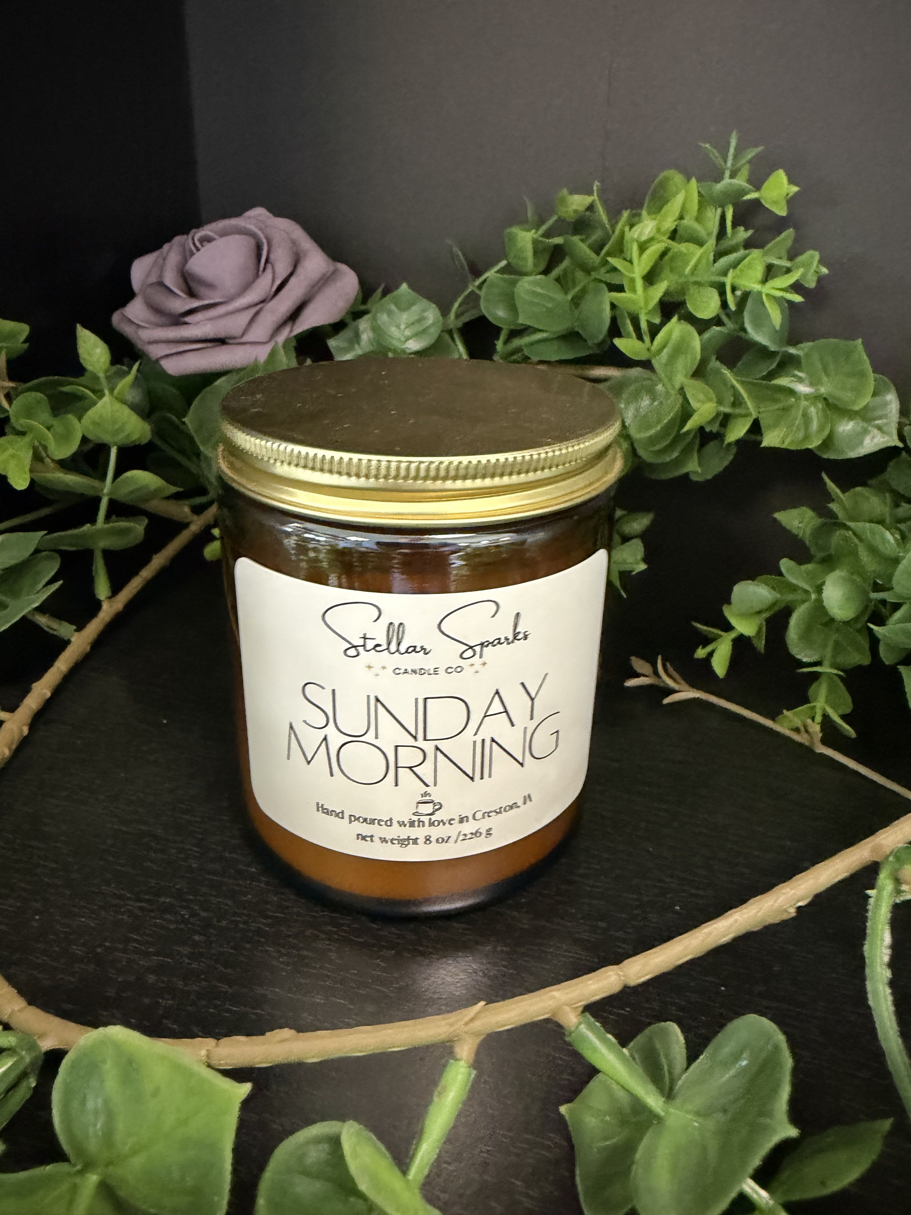 Sunday Morning 8oz Candle - A lavender rose tucked beside faux greenery sets a calm, cozy tone for this 8oz glass jar candle-perfect as a gift for birthdays, housewarmings, or a thoughtful "just because" moment. The product type (flower arrangement paired with candle) and occasion (housewarming, birthday, everyday gift) appear naturally in the presentation, so the person you're thinking of will feel seen by a simple, soothing surprise that smells like a slow Sunday morning.  The candle sits in an amber glass jar with a gold metal lid and a printed "Sunday Morning" label, styled with silk greenery and a single lavender rose for a soft, rustic look. A decorative wreath frames the jar for added charm; color accents include calming greens, pale purple, white, and neutral amber tones. The faux flowers mean no wilting-this styling stays fresh on a mantel, bedside table, or entry console.  Ideal for someone who appreciates low-maintenance décor and gentle, aromatic ambiance, this combination reads as a ready-made gift set that communicates care without fuss. Candle size: 8oz in a glass jar with metal lid; styling includes silk roses, faux greenery, printed label, and decorative wreath.