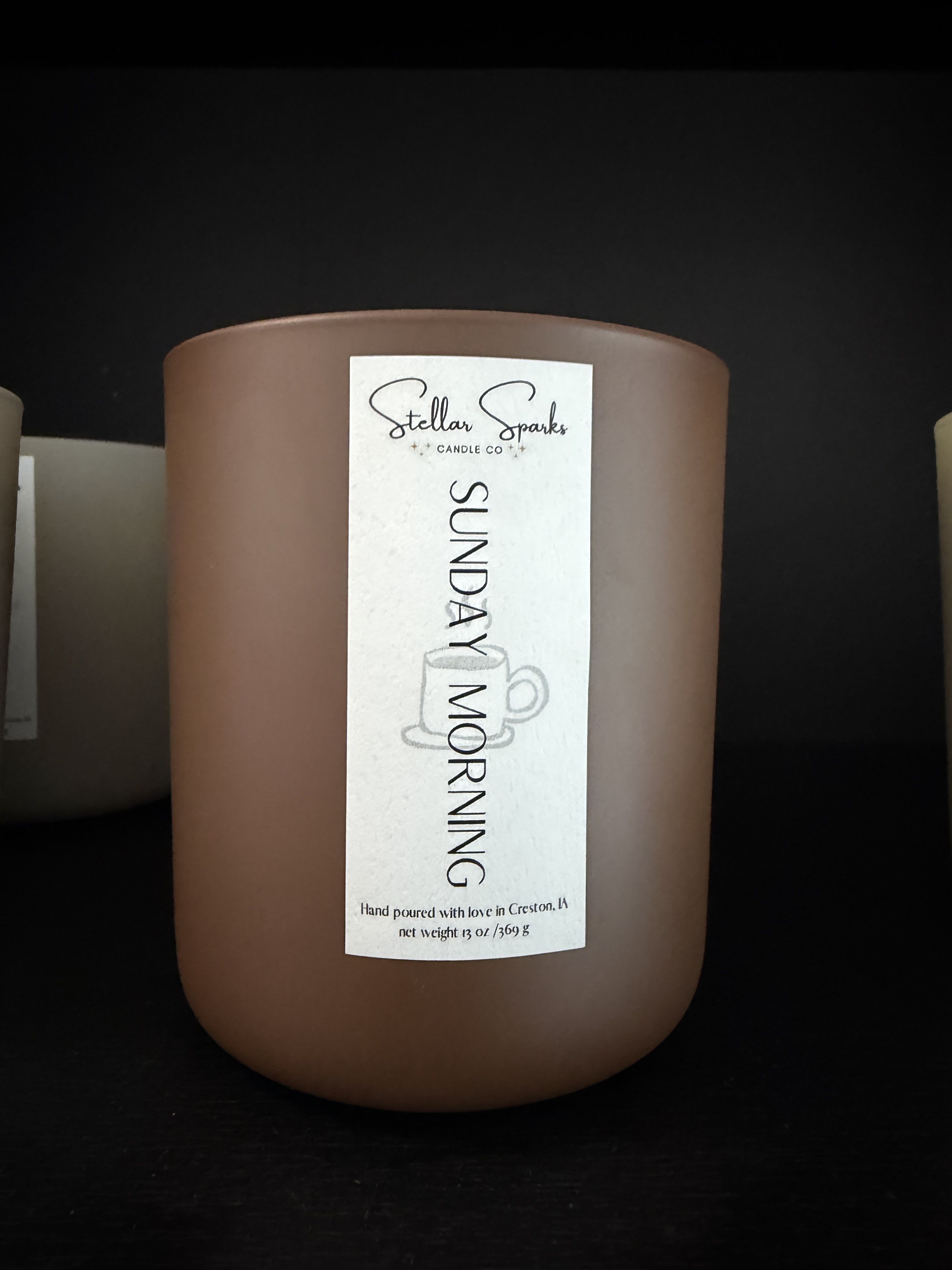 Sunday Morning 13oz Candle - A warm, neutral centerpiece that quietly transforms a room-this 13oz candle jar is designed as a giftable everyday item to bring calm to mornings, birthdays, housewarmings, or a simple "thinking of you" moment. As a home fragrance piece, Sunday Morning reads like an invitation to slow down: the matte brown cylinder, tall white label and subtle cup illustration give it an understated, cozy charm that suits minimalist and rustic interiors alike.  The candle comes in a 13oz matte brown glass jar with a tall white front label featuring the name "Sunday Morning" and the Stellar Sparks Candle Co. branding. Clean black and white graphics keep the look modern and versatile; the compact, cylindrical shape makes it easy to place on a bedside table, kitchen shelf, or living-room mantel. Packaged as a ready-to-gift item, it's an approachable choice for anyone who appreciates quiet, everyday luxury.