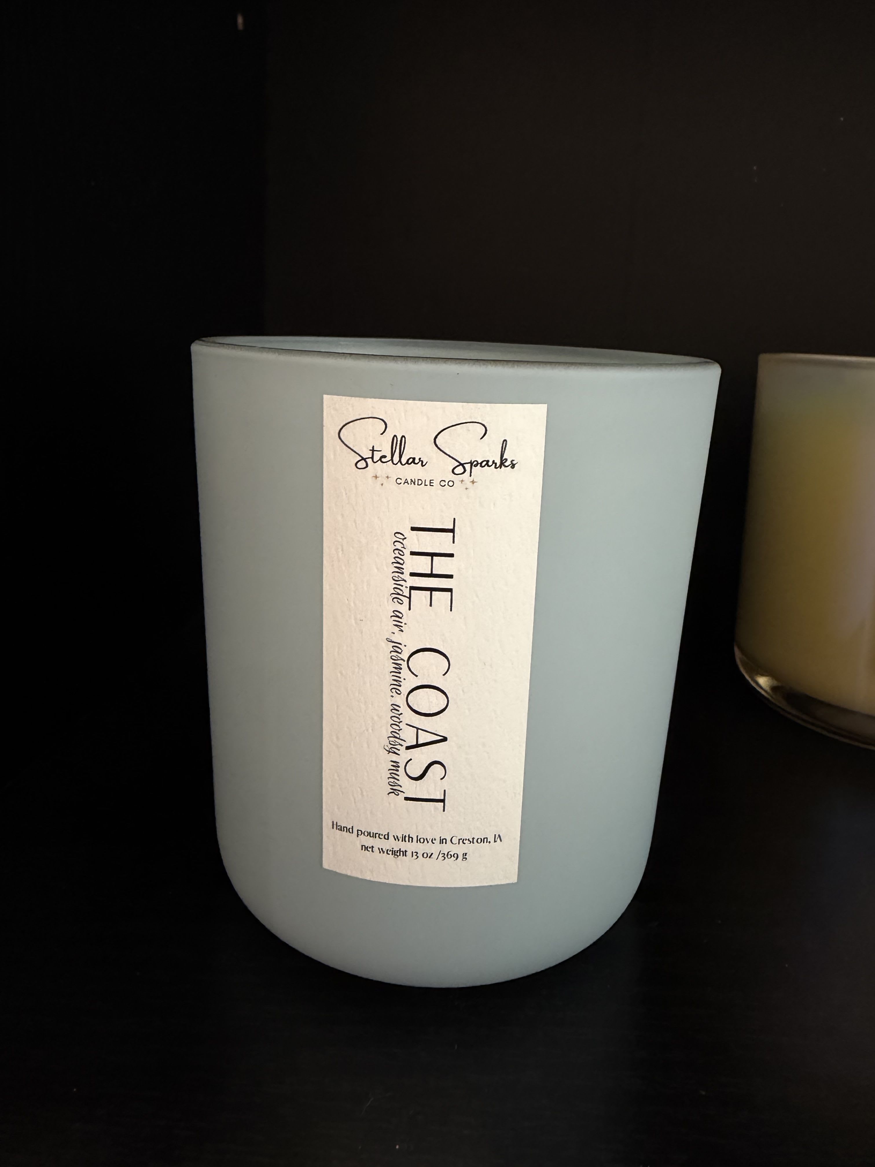 The Coast 13oz Candle - Pale, matte light-blue glass and a clean rectangular paper label give this home fragrance a calm, coastal presence - perfect as a housewarming gift or a thoughtful birthday present for someone who loves serene, beachy decor. The product is a 13oz candle presented in a minimalist glass jar labeled "The Coast," an understated choice for sending a peaceful, spa-like moment to the person you're thinking of.  The candle comes in a large, matte light-blue jar with a tall, dark-ink paper label that reads "The Coast" and notes Stellar Sparks Candle Co., creating a crisp white-and-blue palette with black type for contrast. The smooth glass and muted color make it easy to style on a mantel, bedside table, or bathroom shelf; the neutral graphic label keeps the look modern and gift-ready.  Ideal for housewarmings, hostess gifts, or as a small anniversary or thank-you present, this candle arrives ready to display and enjoy. Size: 13 oz in a glass jar with paper label - burn and care instructions are included on the packaging.