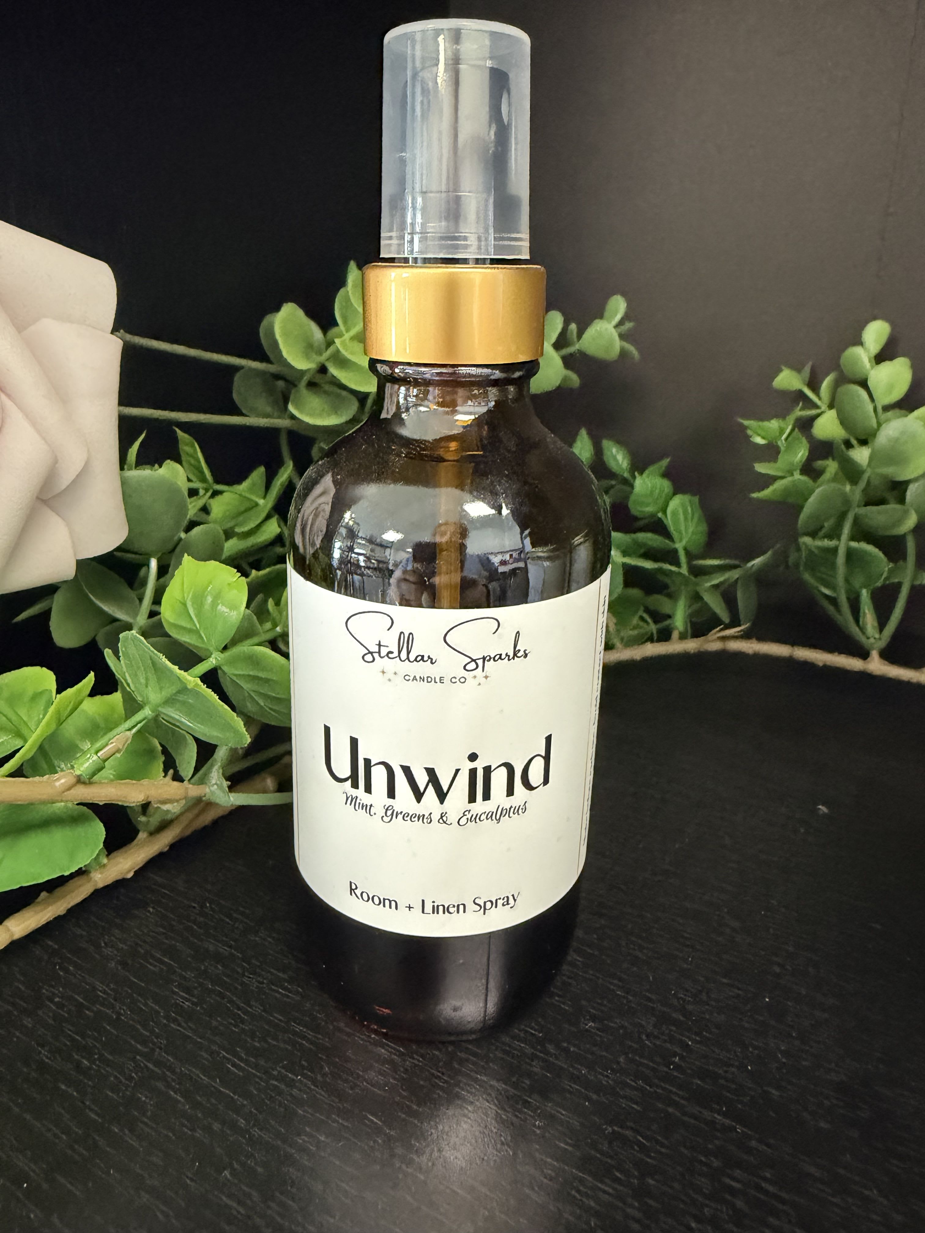 Unwind Room Spray - A cool, spa-like scent that helps someone pause and breathe - this Unwind room spray is a home fragrance spray ideal for gifting as a thoughtful housewarming present or a calming birthday treat. The product type (room and linen spray) and occasion (housewarming, gift) appear naturally up front so the person you're buying for immediately knows this will freshen bedrooms, living rooms, or linens with a soothing, green aroma.  The amber glass bottle with clear cap and plastic spray nozzle is styled simply and elegantly; a white label reads "Unwind" and "Mint, Greens &amp; Eucalyptus," highlighting the clean, botanical fragrance. Visual accents of pale rose and eucalyptus in the styling suggest subtle floral and verdant notes; the bottle's green, white and black tonal palette keeps the look modern and gift-ready. Size and exact volume are shown on the product label.  Perfect for someone who needs a moment of calm, this spray makes a low-fuss addition to a self-care kit, bedside table, or entryway; it's the kind of small luxury that helps the recipient feel noticed and relaxed. Spray lightly on linens or into the air for an instant refresh - store upright and avoid direct heat.
