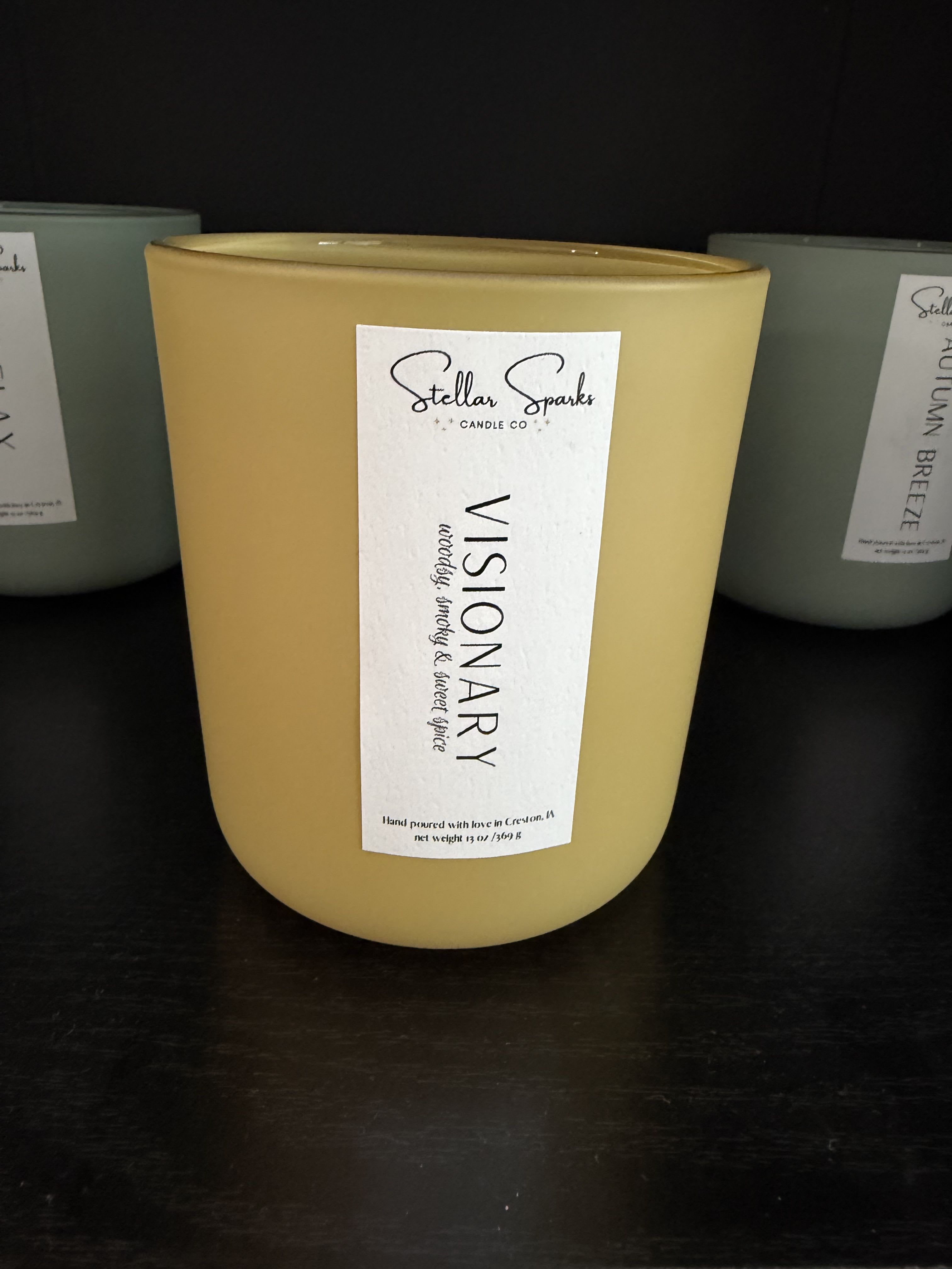 Visionary 13oz Candle - A calm, minimalist scented candle designed to bring a warm, cozy glow to any room - the perfect home-fragrance gift for a housewarming or a thoughtful birthday present. This flower-arrangement product listing highlights the Visionary 13oz candle as a companion gift to floral bouquets or a standalone present for someone who loves quiet, layered scent moments.  Hand-poured into a smooth beige ceramic jar with a tall white paper label reading "Visionary," the 13oz candle blends clean lines and neutral tones-yellow, white, and black appear in product styling-so it complements both bright bouquets and soft, modern interiors. The simple packaging and ceramic vessel make it easy to restyle on a mantel, bedside table, or entry console; the jar can be reused once the candle is finished. Order as an add-on to your arrangement or alone for immediate delivery; burn responsibly and trim the wick to 1/4" before each use for an even, long-lasting fragrance experience.