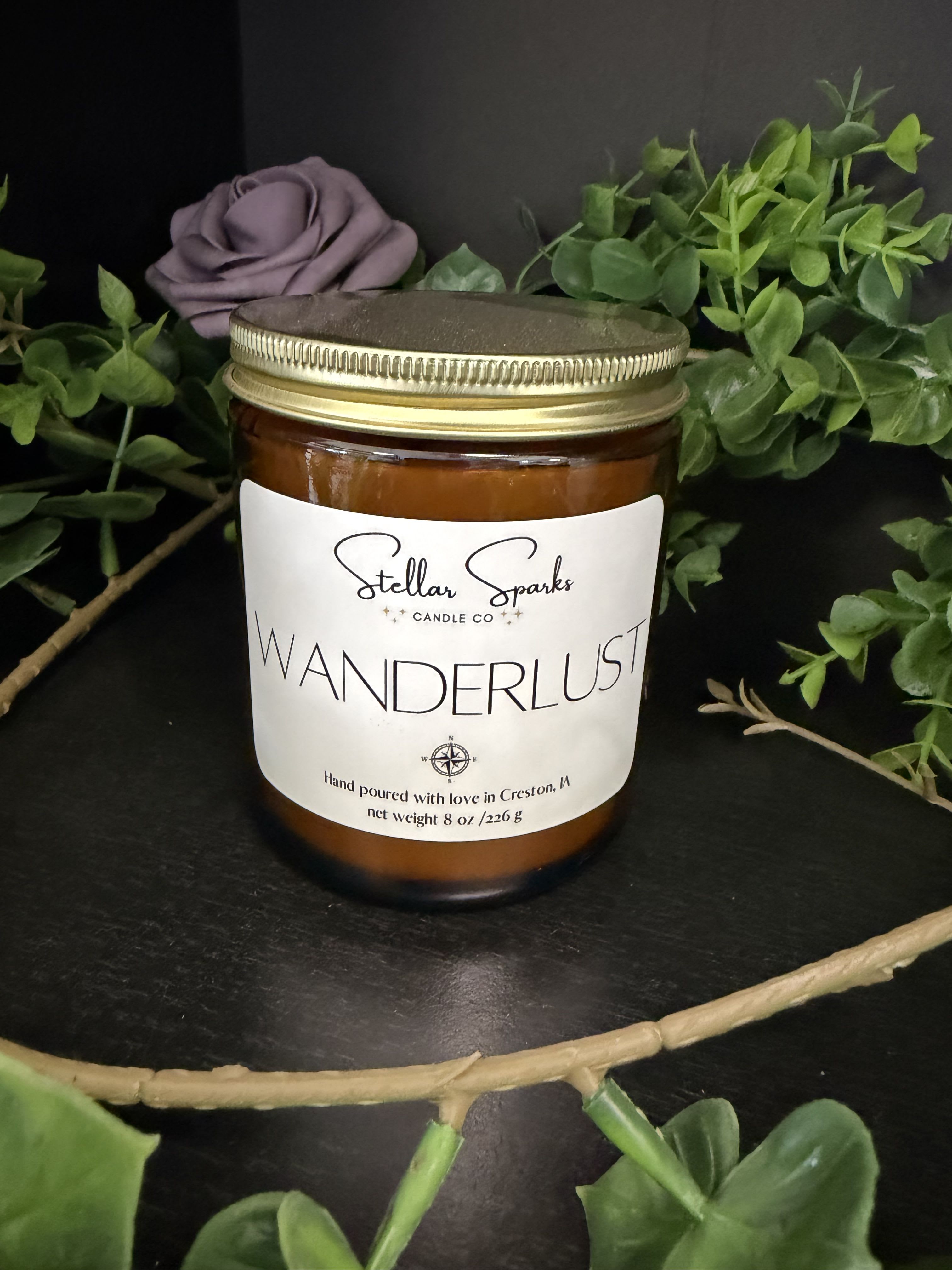 Wanderlust 8oz Candle - A moody, artisanal 8oz jar candle that reads like a little travel memory in wax-perfect as a gift or a housewarming accent. This candle is presented as a home fragrance gift that complements floral-scented moments and makes a thoughtful present for someone who loves cozy, rustic décor or a wanderlust spirit.  The candle comes in an amber glass jar with a gold metal lid and a clean white front label printed with "WANDERLUST" and "Stellar Sparks Candle Co," giving it an upscale, handmade feel. Styled here with faux greenery and a muted purple bloom, the design leans green, white, black and warm amber tones-an easy fit on a mantel, bedside table, or entry console. The 8oz size is convenient and burn-friendly for small spaces.  Give this as a thoughtful housewarming, birthday, or "thinking of you" gift that suggests escape and warmth-she'll notice the thoughtful presentation as much as the fragrance. For best results, place on a heat-resistant surface, trim the wick to 1/4" before each burn, and enjoy steady, even fragrance across multiple hours.