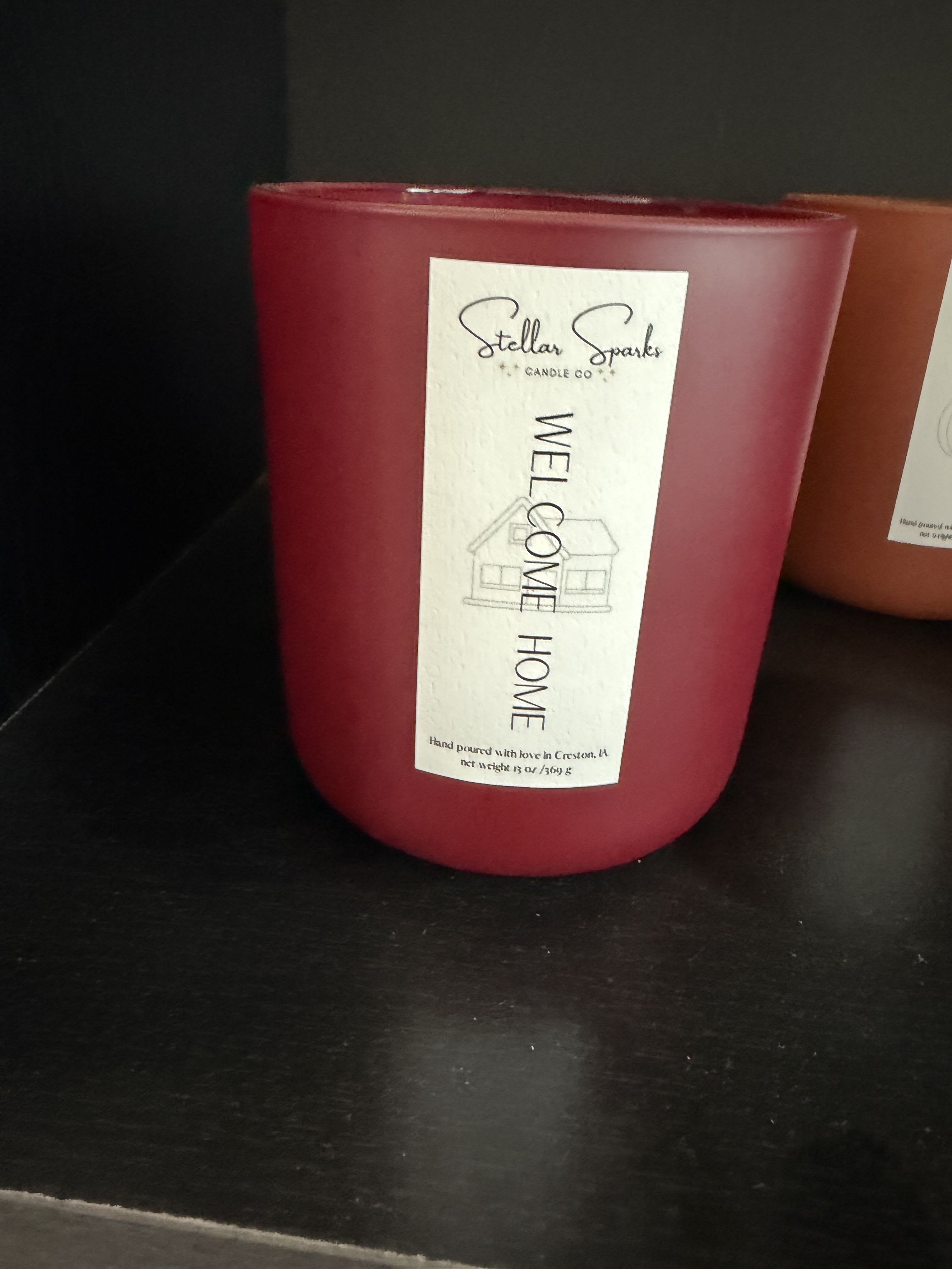 Welcome Home 13oz Candle - A deep red, 13oz welcome-home candle that greets a new space with cozy warmth - perfect as a housewarming gift or a thoughtful present for someone settling into a new apartment. The arrangement-style presentation is simple and inviting: the candle itself reads "Welcome Home" on a tall rectangular label with a minimal house illustration, making it an immediate, sentiment-forward choice for celebrating a move or welcoming someone back.  Poured into a frosted glass jar, the rich red wax contrasts with the clean white and orange accents on the printed label for a modern, homey look that complements most decor. The jar's smooth frosted finish and the neat label give it a polished, gift-ready appearance; the 13oz size provides hours of burn time while staying compact enough for a shelf, entry table, or bedside. Presented alone or paired with seasonal flowers, this candle makes the recipient feel seen and settled - an uncomplicated way to say "congratulations" or "thinking of you" for new homes, open houses, or cozy evenings in.