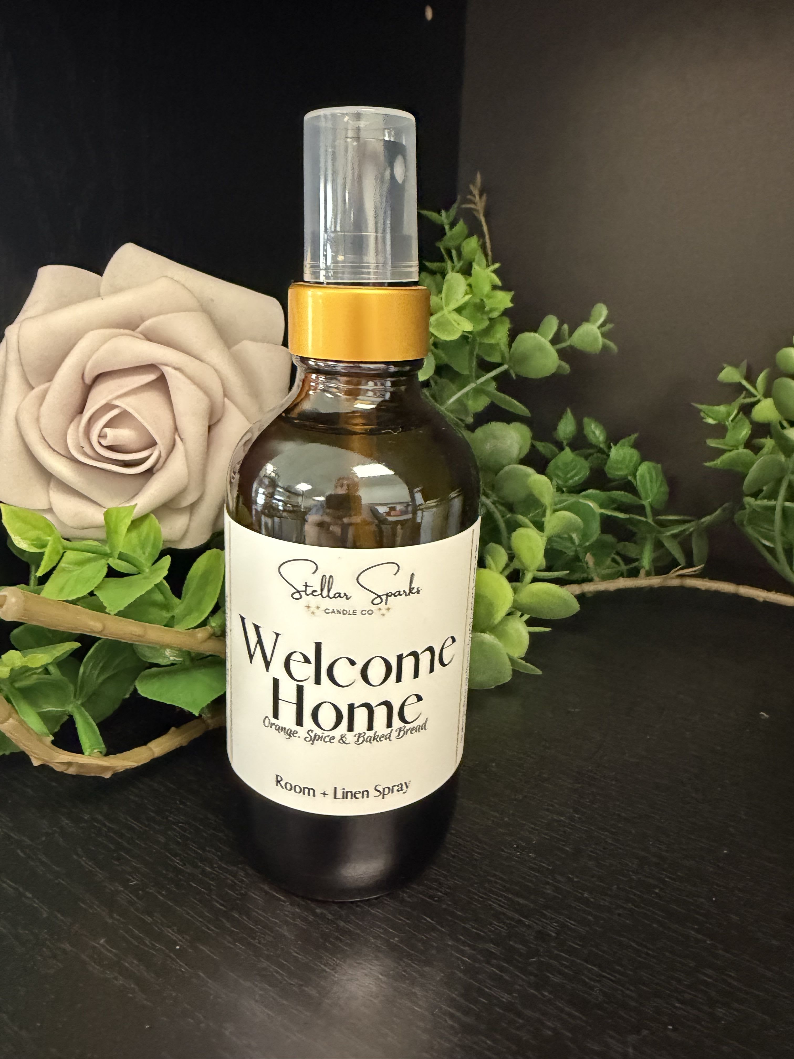 Welcome Home Room Spray - An amber glass room spray crowned with a cream "Welcome Home" label-this welcome home room spray is the perfect housewarming or everyday gift to make someone's space feel intentionally cozy. The presentation pairs the spray bottle with a soft beige rose and bright green faux foliage so the person you're celebrating will see and smell a thoughtful, decorative touch right away.  The spray comes in a recyclable amber glass bottle with a clear cap and plastic spray nozzle, finished with a large, elegant product label. Colors in the styling are warm neutrals and fresh green accents that suit entryways, living rooms, or bedside tables; the included floral accent is a beige rose set against vivid faux greenery for a natural, lived-in look.  Give this to a new homeowner, someone settling into a new apartment, or the friend who could use a little welcome-she'll feel immediately seen every time the room fills with scent. Size and exact fragrance notes are on the label; store upright, keep out of direct sunlight, and mist lightly over linens or into room air as desired.
