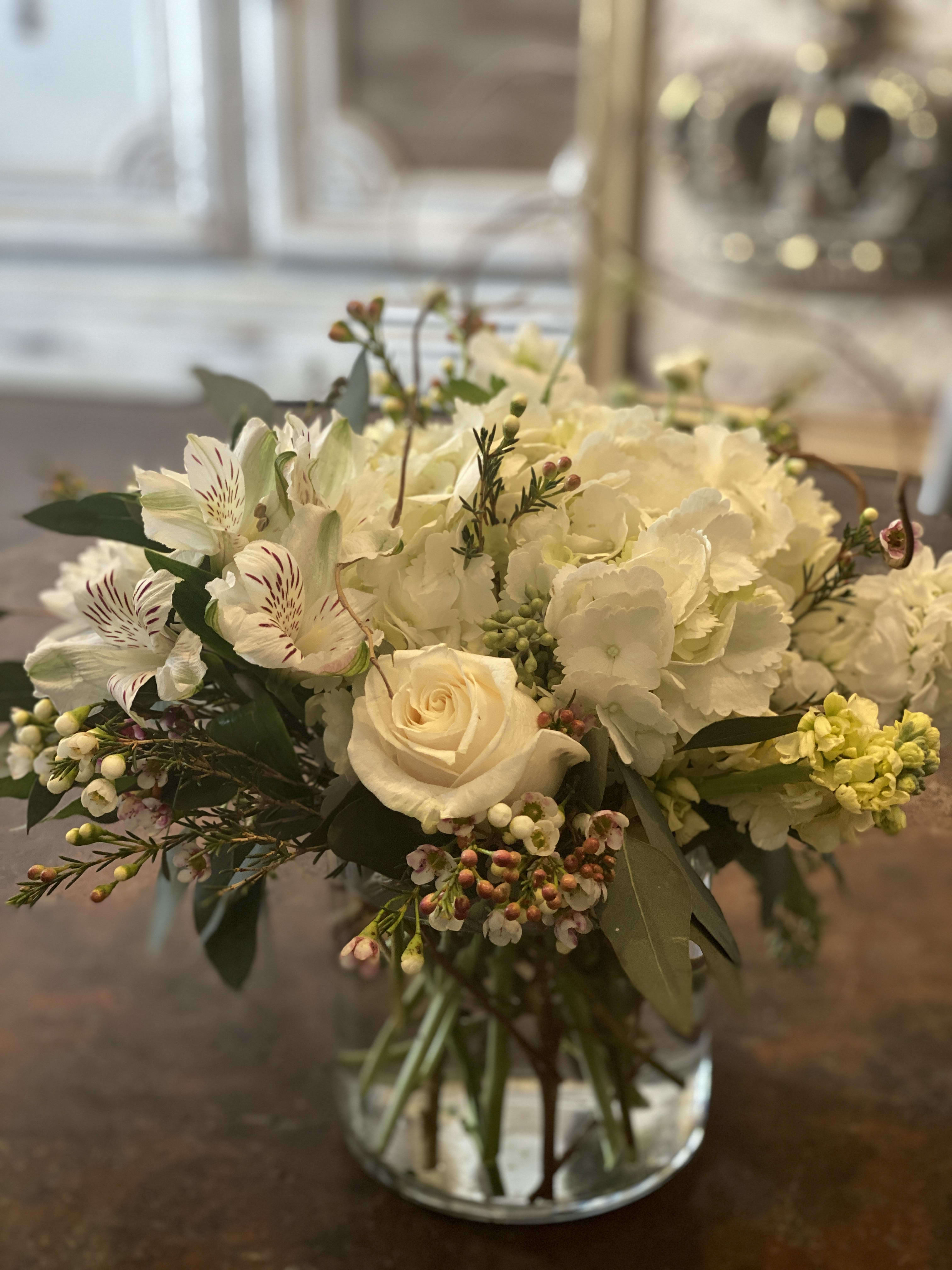 Blooms Pretty - White roses, hydrangeas, greens and more are arranged in a short cylinder glass vase. This bouquet will look perfect on a coffee table or kitchen counter and will enhance both a modern or traditional look. 