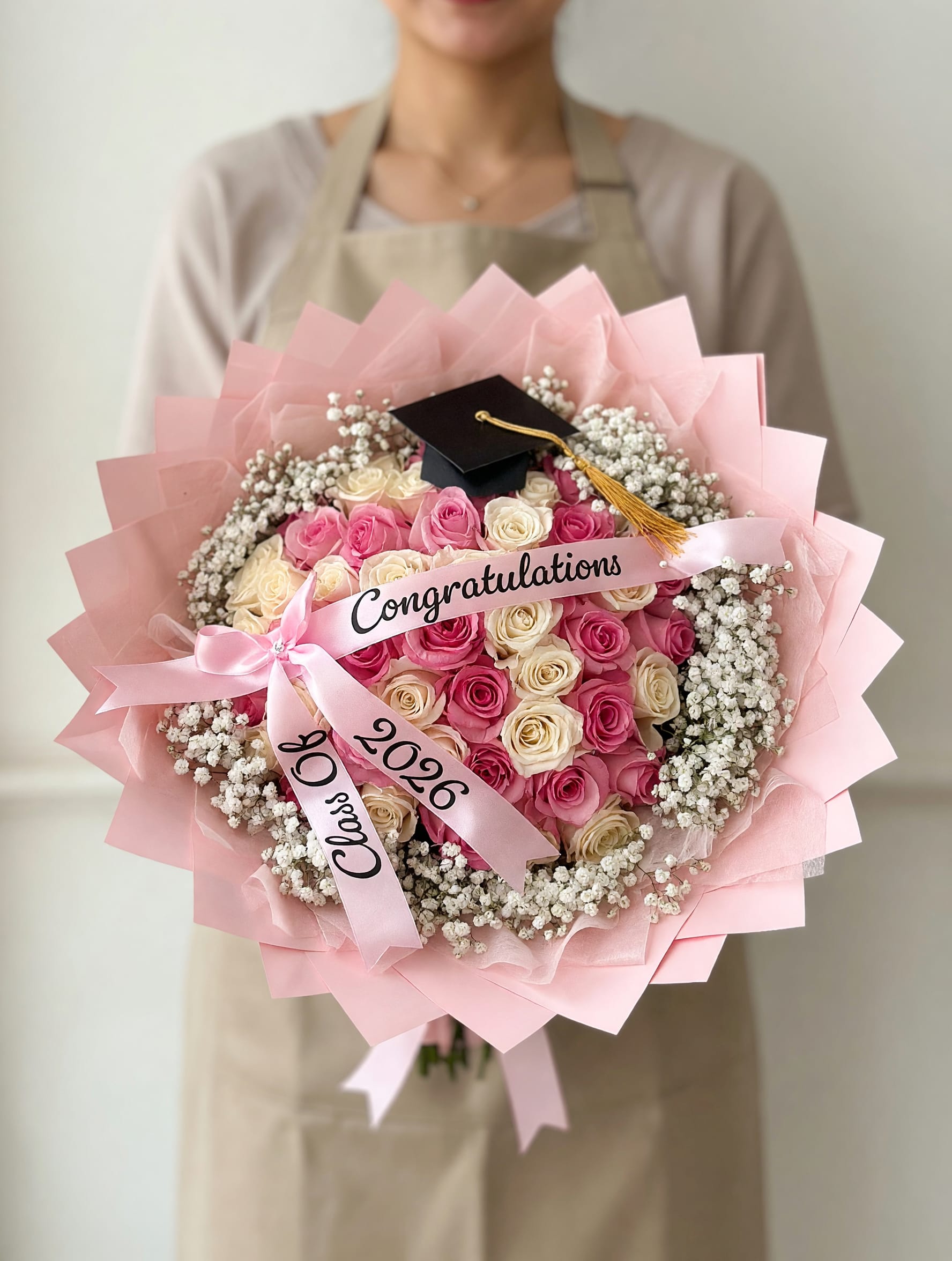 50 Roses Pink &amp; White Graduation Bouquet - A lush cascade of fifty pink and cream roses arranged into a generous hand-tied graduation bouquet-this arrangement announces celebration the moment she sees it. Perfect as a graduation bouquet or congratulatory gift, the tightly packed roses create a soft, romantic cloud of color while clusters of white baby's breath ring the blooms for a fresh, airy contrast. A small black graduation cap tucked near the top and a satin ribbon printed "Congratulations" with "Class of 2026" on the tails make the occasion unmistakable.  Wrapped in soft pink paper and finished with a matching pink ribbon, the bouquet arrives ready to present and makes the recipient feel proudly celebrated and seen. The combination of pink, cream, and white offers a feminine, cheerful palette that suits new graduates, sisters, daughters, or friends stepping into their next chapter. Hand-tied for easy handling, this sizable bouquet makes a memorable photo moment and the kind of gift that keeps the accomplishment front and center.