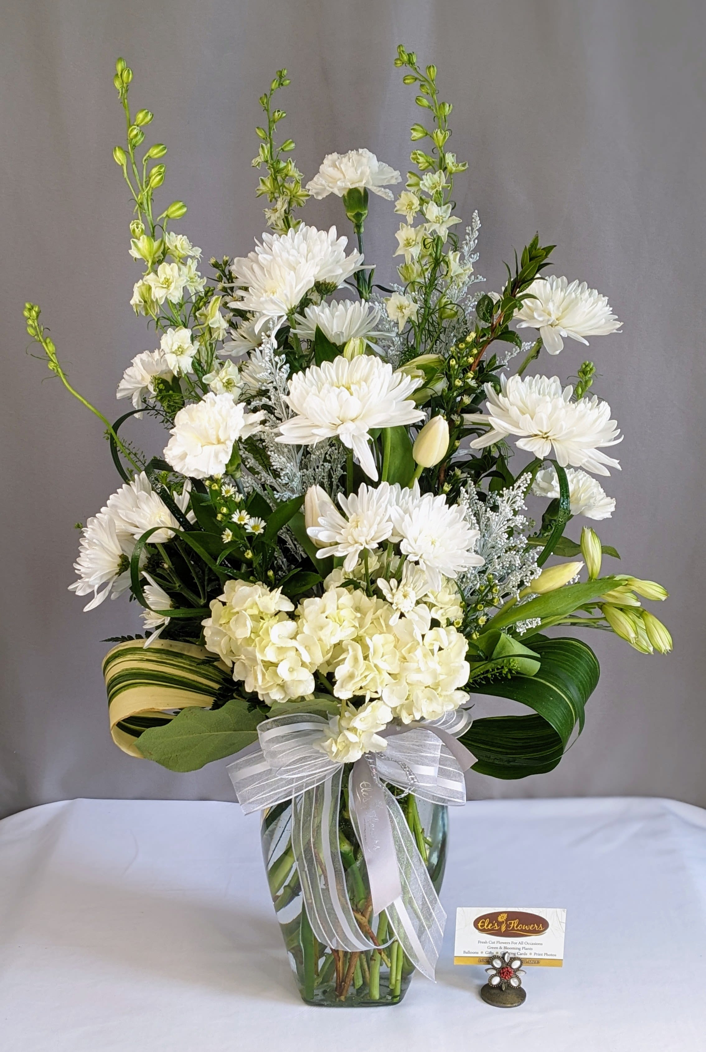 Serene White - Sympathy Arrangement - As peaceful and beautiful as a starry night, this pure white funeral bouquet is a touching way to remember a truly special person. Send this classic arrangement to comfort the newly bereaved at the funeral. Fresh white flowers are accompanied by lush greenery and accents to create a stunning bouquet of remembrance.    The designs we create are custom and the blooms and containers will vary. We use the freshest product available to create the perfect floral combination for the recipient! If you have any special requests, please add those in the "special instructions" portion of your order to help us dial in your vision. Otherwise, our skilled designers, Brett &amp; Sara, will take it from here!   *Please note that the photos are examples only – floral selection and containers will vary.*  Standard Size is an excellent option for a kitchen island. Approximate. Dimensions: ~20”H x 12”W  Deluxe Size is a great and showy, grand size for a larger kitchen table. Approximate. Dimensions: ~25"H x 18"W  Premium Size is an extravagant and luxurious display for a funeral service or celebration. Approximate Dimensions: ~30"H x 22"W  