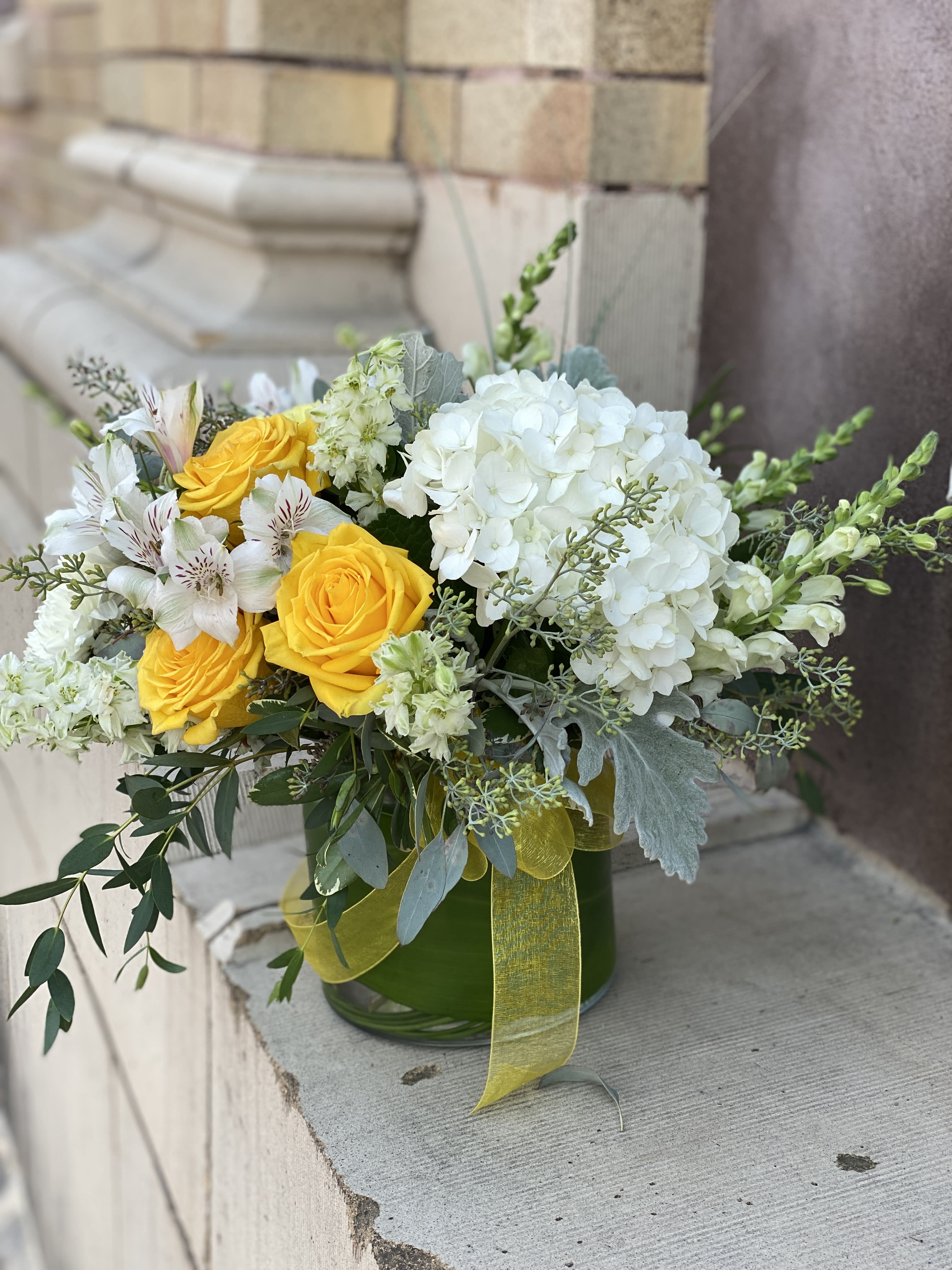 Low glass vase of yellow roses, white hydrangeas, and white blooms with a yellow ribbon.