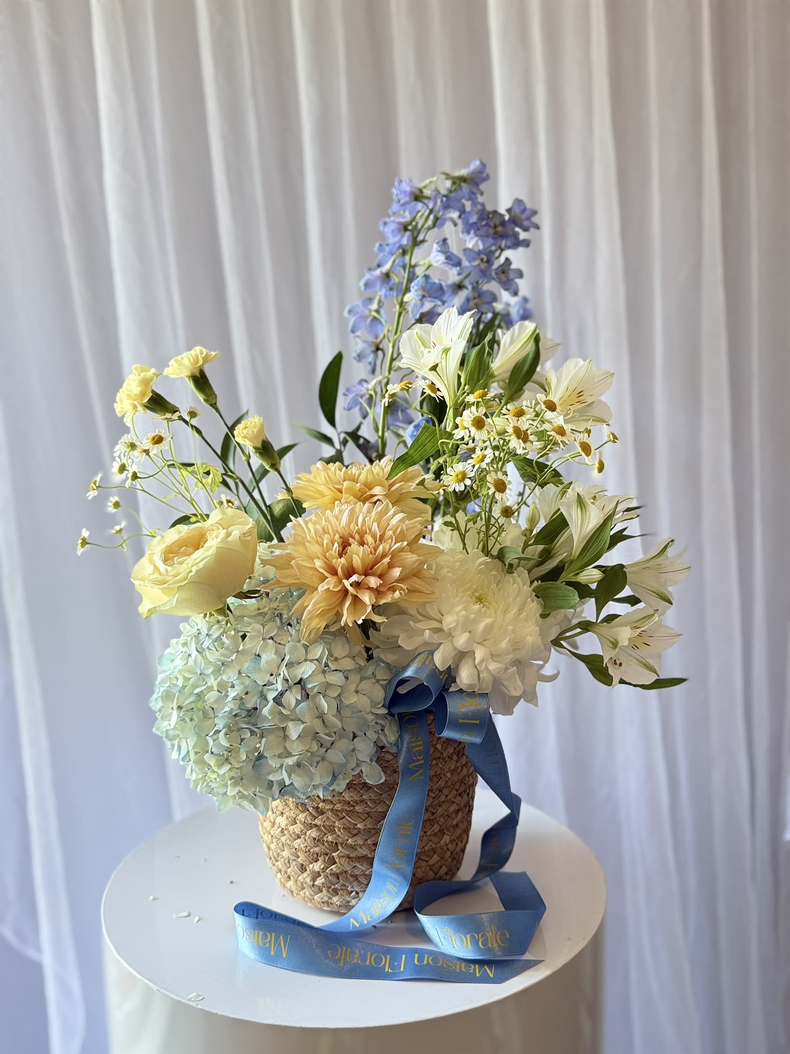 Côte d'Azur - Côte d'Azur is a sun-drenched arrangement inspired by the colours of a Mediterranean summer — the soft gold of afternoon light, the cool blue of a cloudless horizon, and the quiet white of sea-washed linen. Hand-arranged in a woven seagrass basket, this piece brings together café au lait dahlias, yellow garden roses, powder blue delphinium, white alstroemeria, blush hydrangea, and delicate chamomile — each stem chosen for its texture, its colour, and the feeling it brings to the room.