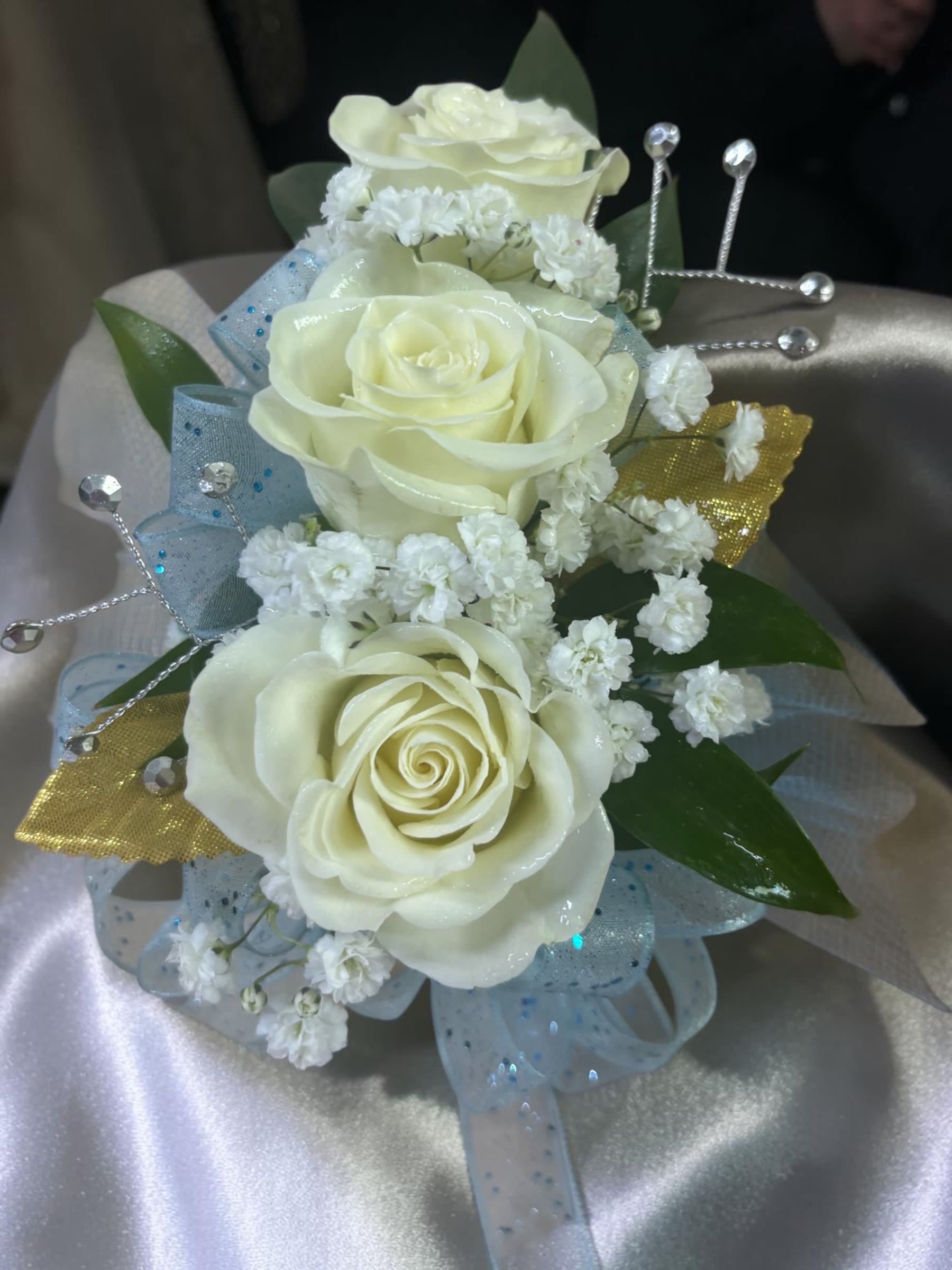 Golden Sky Wrist Corsage - Golden Sky Wrist Corsage combines classic white spray roses with soft light blue ribbon, shimmering gold metallic leaves, rhinestone accents, accent flowers, and fresh greenery for an elegant and unforgettable prom look. Available in Standard, Deluxe, and Premium sizes to fit a variety of styles and budgets.  Standard features 3 white spray roses, Deluxe features 4 white spray roses, and Premium features 5 white spray roses for a fuller presentation.  Looking for the matching boutonnière? Find our Golden Sky Boutonnière in the Boutonnière category. Looking for a matching corsage? Find our Golden Sky Wrist Corsage in the Corsage category.  Designed for prom, homecoming, winter formal, and other school dances, this design offers a timeless combination of elegance, sparkle, and fresh floral beauty.  Accent flowers and decorative elements may vary based on availability while maintaining the overall style, color palette, and value of the design. Each piece is handcrafted, and no two designs for a local dance are exactly alike.