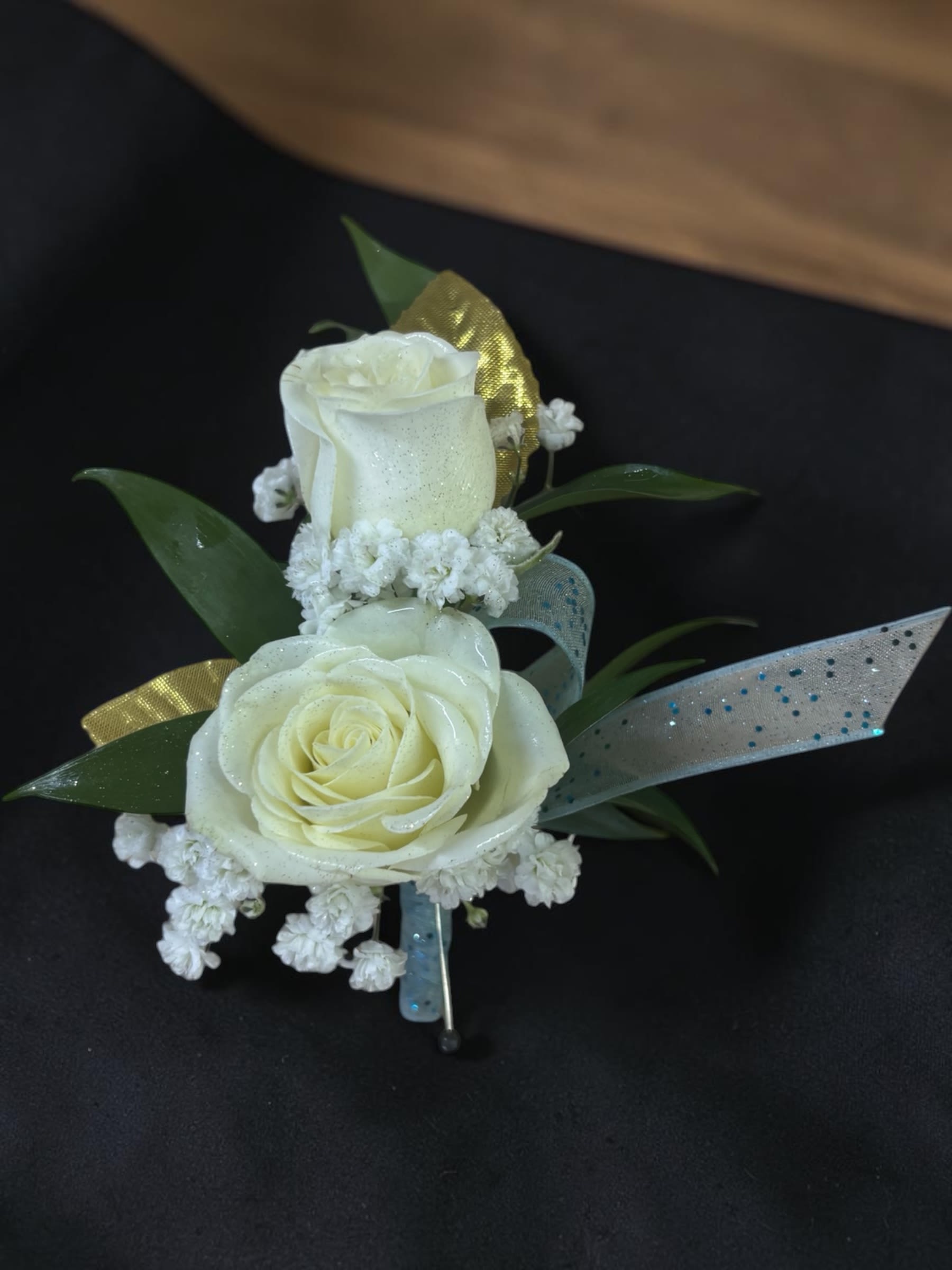 Golden Sky Boutonniere - MAGNET ATTACHMENT INCLUDED. WARNING: WEARERS WITH PACEMAKERS, DEFIBRILLATORS, INSULIN PUMPS, HEARING DEVICES, OR OTHER MEDICAL DEVICES MUST REQUEST A PIN ATTACHMENT WHEN ORDERING.  Golden Sky Boutonnière features two white spray roses accented with light blue ribbon, gold metallic leaves, accent flowers, and fresh greenery for a classic and elegant look. Designed for prom, homecoming, winter formal, and other school dances, this boutonnière provides the perfect finishing touch for any formal outfit.  Looking for the matching corsage? Find our Golden Sky Wrist Corsage in the Corsage category.  Accent flowers and decorative elements may vary based on availability while maintaining the overall style, color palette, and value of the design. Each piece is handcrafted, and no two designs for a local dance are exactly alike.