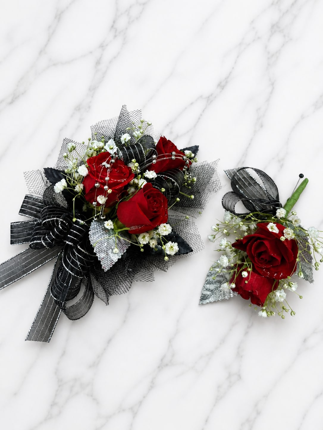 Putting on the Ritz Corsage - Putting on the Ritz Wrist Corsage combines rich red spray roses with black and silver accents, ribbon, rhinestone accents, accent flowers, and fresh greenery for a dramatic and elegant formal look. Inspired by classic black-tie style, this wrist corsage is designed to make a bold statement at prom, homecoming, winter formal, and other school dances.  Choose from Standard, Deluxe, or Premium options based on the number of roses in the corsage. Standard features 3 red spray roses, Deluxe features 4 red spray roses, and Premium features 5 red spray roses for a fuller presentation.  Corsage Only. Looking for the matching boutonnière? Find our Puttin’ on the Ritz Boutonnière in the Boutonnière category. Find the set in our Prom category.  The striking combination of red roses, black ribbon, silver accents, and sparkling rhinestones creates a timeless design that is both sophisticated and unforgettable.  Accent flowers and decorative elements may vary based on availability while maintaining the overall style, color palette, and value of the design. Each piece is handcrafted, and no two designs for a local dance are exactly alike.