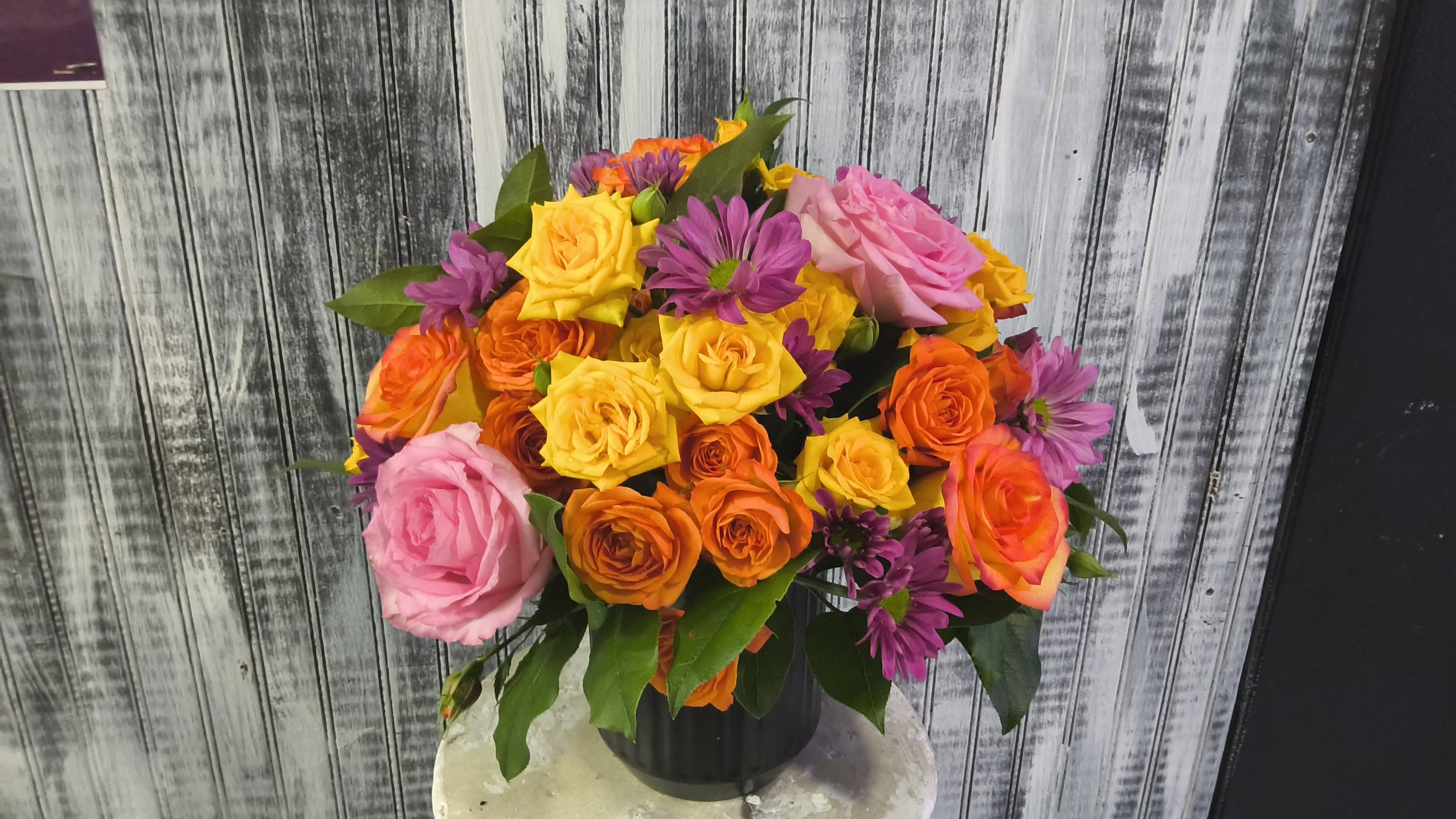 Blossoming sunshine - A lively burst of color-this flower arrangement is a cheerful birthday bouquet or congratulations gift that will make the person you're thinking of feel genuinely celebrated. The composition centers on abundant roses in pink, yellow, and orange, with purple daisy-like blooms threaded throughout to add playful texture and contrast. Paired with a sleek black vase, the bright palette reads joyful and modern rather than formal.  Packed full for a generous presentation, the arrangement's rounded rose heads sit close together for a dense, garden-fresh look while the smaller purple daisies peek between petals to catch the eye. The black vase grounds the vivid hues and suits both casual and contemporary settings. Perfect for birthdays, congratulations, or simply to brighten someone's day-this is the kind of bouquet that delivers an unmistakable "you matter" message.
