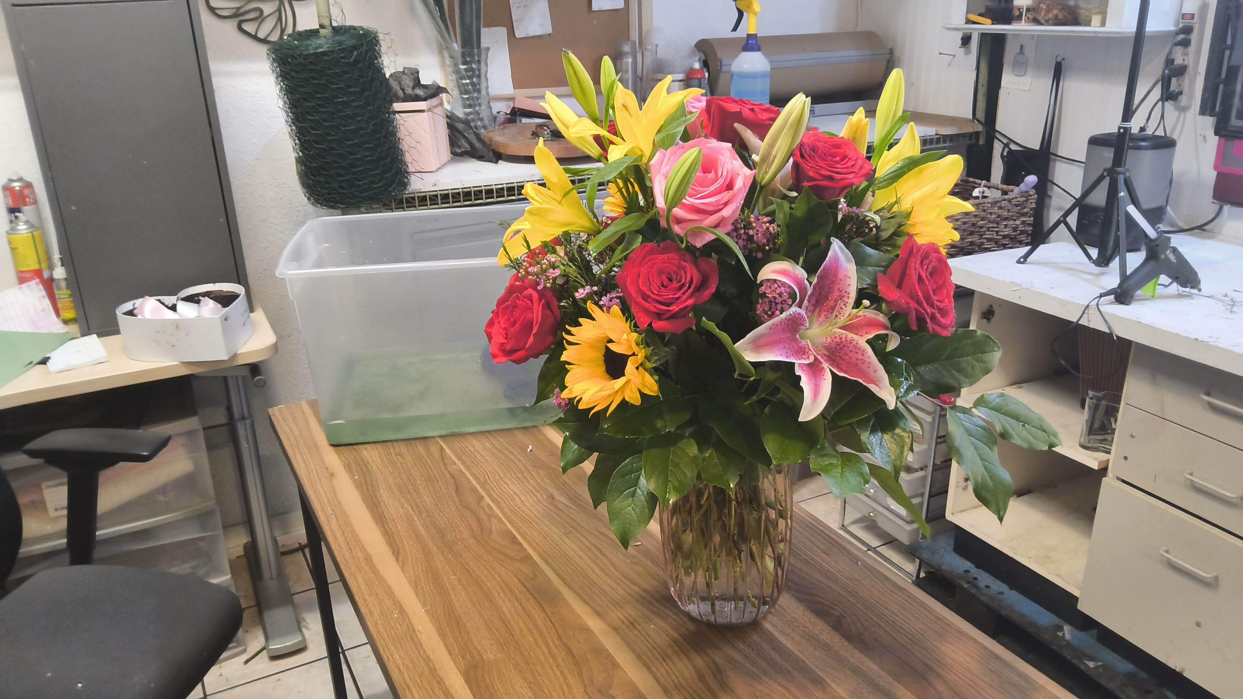Golden glow bouquet  - Bright yellow lilies and sunflowers glow alongside pink and red roses in a full, rounded bouquet-an uplifting birthday bouquet or thank-you gift that reads as joyful and celebratory. The warm mix of colors against rich green foliage creates a lively contrast that makes the recipient feel seen and celebrated, perfect for birthdays, expressions of gratitude, or just because moments.  This arrangement features layered blooms of roses, lilies, sunflowers, and complementary filler flowers arranged in a clear glass vase for a clean, classic presentation. The design is lush and textured-soft rose petals, bold sunflower faces, and the trumpet shape of lilies combine to give depth and movement while dark greenery frames each bloom. Suitable for same-week delivery, the vase arrangement arrives ready to display; change the water every couple of days and trim stems to keep the flowers fresh longer.