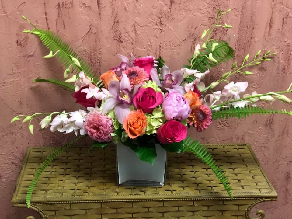 Keep Her Wild - Type of Flowers: Green Hydrangeas, Seasonal Pink Peonies, Seasonal Pink Dahlias, Lavender Cymbidium Orchids, Coral and Hot Pink Roses, Pink Dendrobium Orchids, and Fern in a 6" x 6" white, crystal, square vase. Availability: Peonies available November to mid-August, Dahlias available mid-summer to Autumn Substitute Available: Yes Design View: Front Symmetric View Photo shown: Regular 