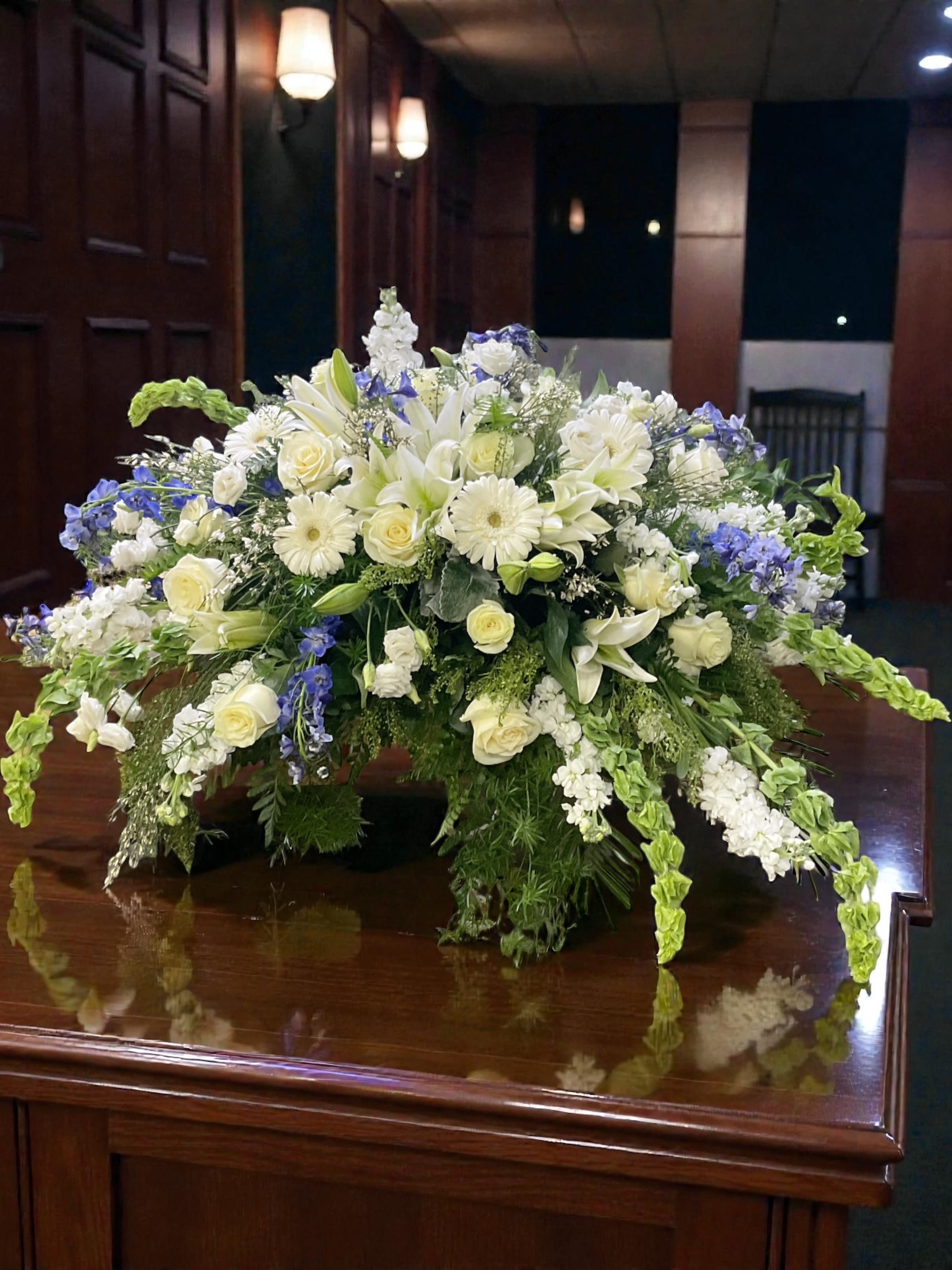 Beside the River Casket Spray - Beside the River is a serene white and blue casket spray designed to honor a life with quiet grace and dignity. Soft white roses, lilies, and seasonal florals are layered with cool blue accents, creating a gentle, flowing composition that feels peaceful without being cold.  The design has a natural movement across the casket, offering a sense of calm presence at the service while still carrying enough fullness to feel complete and intentional. It’s the kind of tribute that doesn’t demand attention—but holds it.  A meaningful choice for casket services, memorial services, or celebrating a loved one’s life with softness, balance, and understated beauty.  Available for casket flower delivery through Aspen Tower Florist, serving Greensburg and Shelbyville along with Rushville, Batesville, Westport, Oldenburg, St. Paul, Waldron, and Milroy. 