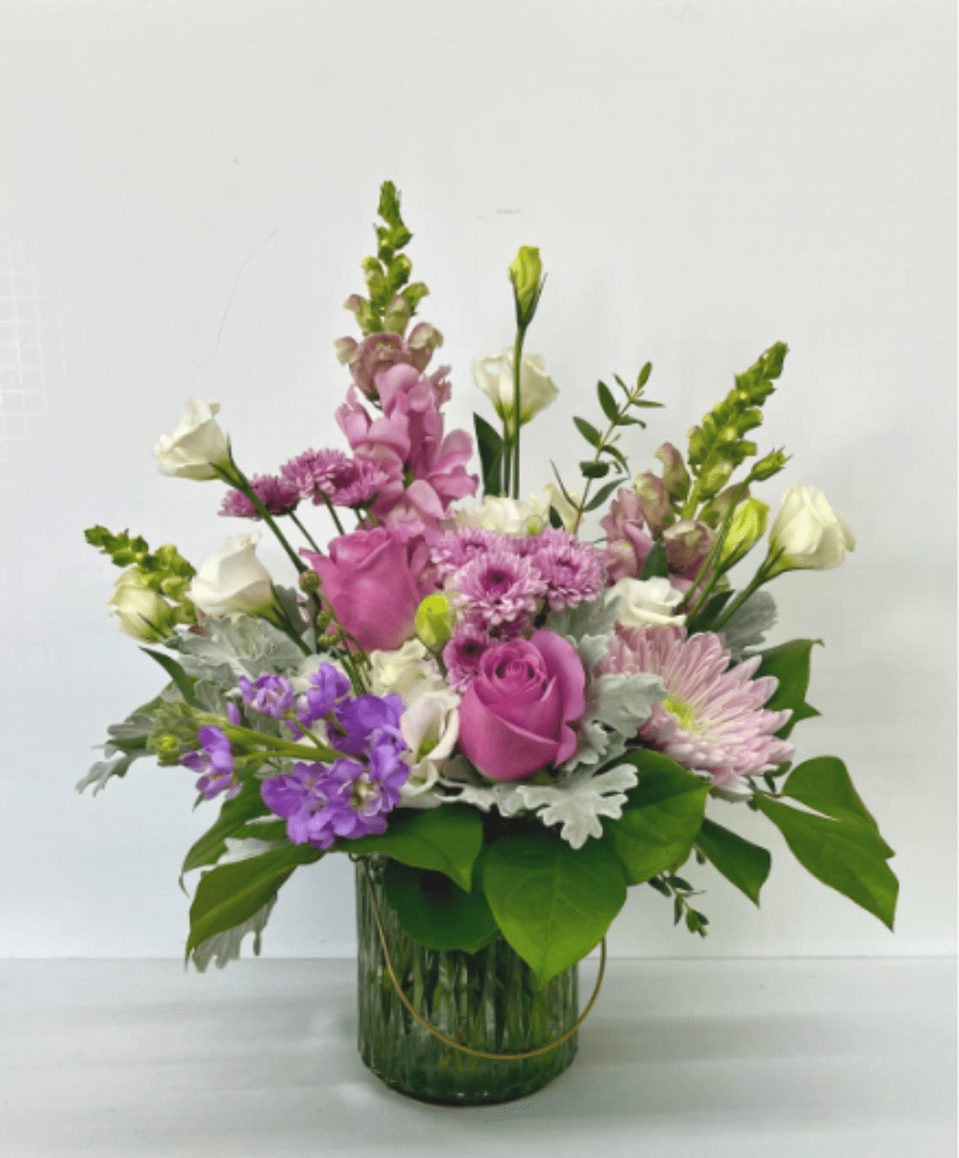 Lantern of Love - A stunning garden-style floral arrangement designed in soft shades of lavender and rich purple, beautifully accented with touches of crisp white. This romantic design features a lush mix of snapdragons, roses, stock, mums, lisianthus, and fresh greenery, artfully arranged in a modern glass vase for a perfect balance of elegance and natural charm.  With its airy, organic style and soothing color palette, Lantern of Love is ideal for birthdays, anniversaries, sympathy gestures, or simply brightening someone’s day. Each arrangement is handcrafted to highlight seasonal blooms and fresh textures, making every piece unique.  Delivery Available In: Smyrna, Marietta, Mableton, Austell, and Atlanta.