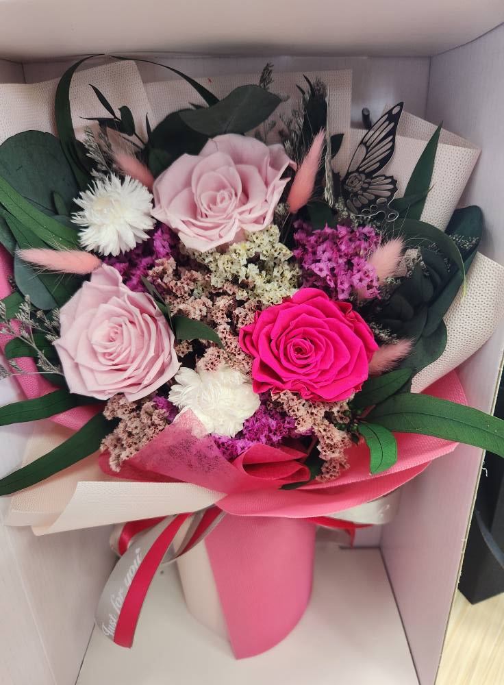 Preserved Rose Bouquet - Preserved roses-pale blush, vivid hot pink, and soft lavender-sit at the heart of this preserved rose bouquet, creating an immediate romantic and cheerful impression perfect for a birthday bouquet, anniversary roses, or a heartfelt thank-you. The playful butterfly accent and layered pink wrapping give the arrangement a feminine, gift-ready look that will make the person you're thinking of feel truly celebrated the moment they open the box.  Surrounding the focal roses are clusters of small filler blooms in white, lavender, and dusty pink, with dark green foliage to add depth and contrast. The arrangement arrives tucked into a presentation gift box with decorative wrapping paper, ribbon, and a delicate butterfly decoration-everything positioned for an elegant reveal and easy gifting.  Ideal for everyday gifting or marking special moments, this preserved bouquet offers lasting beauty without the need for daily care; keep it out of direct sunlight and away from moisture to preserve color and texture. Available in a single polished presentation size that's ready to place on a bedside table, desk, or mantel, it's a thoughtful way to say "I'm thinking of you" with enduring charm.
