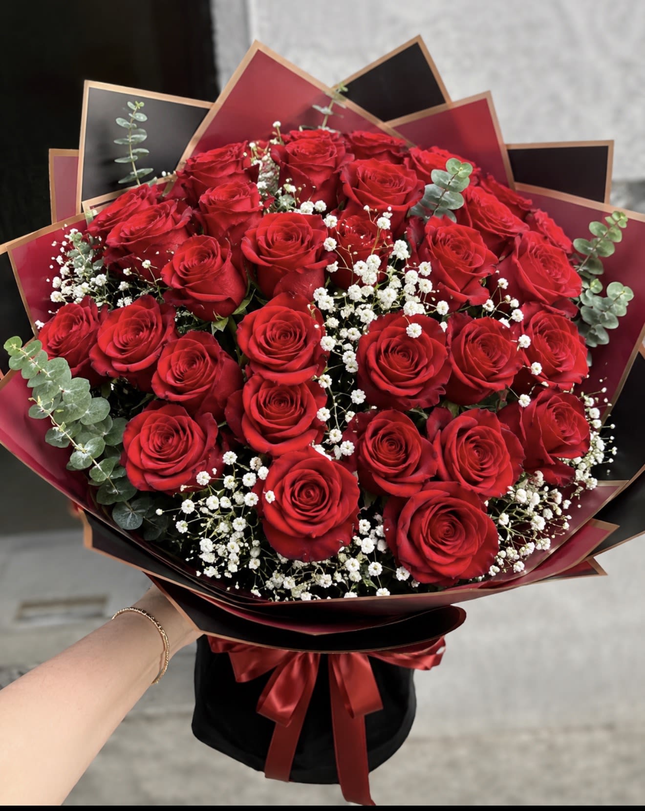 BUCHON - A dense cluster of red roses arranged with delicate white filler and silvery eucalyptus creates a bold, romantic bouquet perfect for an anniversary, birthday, or any love-and-romance moment. This flower arrangement reads dramatic and luxurious thanks to its red-and-black palette-an immediate visual that tells the person you're thinking of them with confidence and warmth.  Wrapped in layered black and burgundy decorative paper and finished with a satin ribbon, the presentation feels polished and intentional. The roses are closely set for a lush, velvety look while the small white blooms and eucalyptus add texture, contrast, and a soft, fragrant lift. Sized as an everyday gift bouquet, it's ideal for celebrating someone special, saying thank you, or marking a meaningful milestone.  Care is simple: keep stems in fresh water, recut the stems before placing in a vase, and trim foliage below the waterline to extend vase life. Choose this arrangement when you want your person to feel seen-romantic, appreciated, and a little bit indulged.