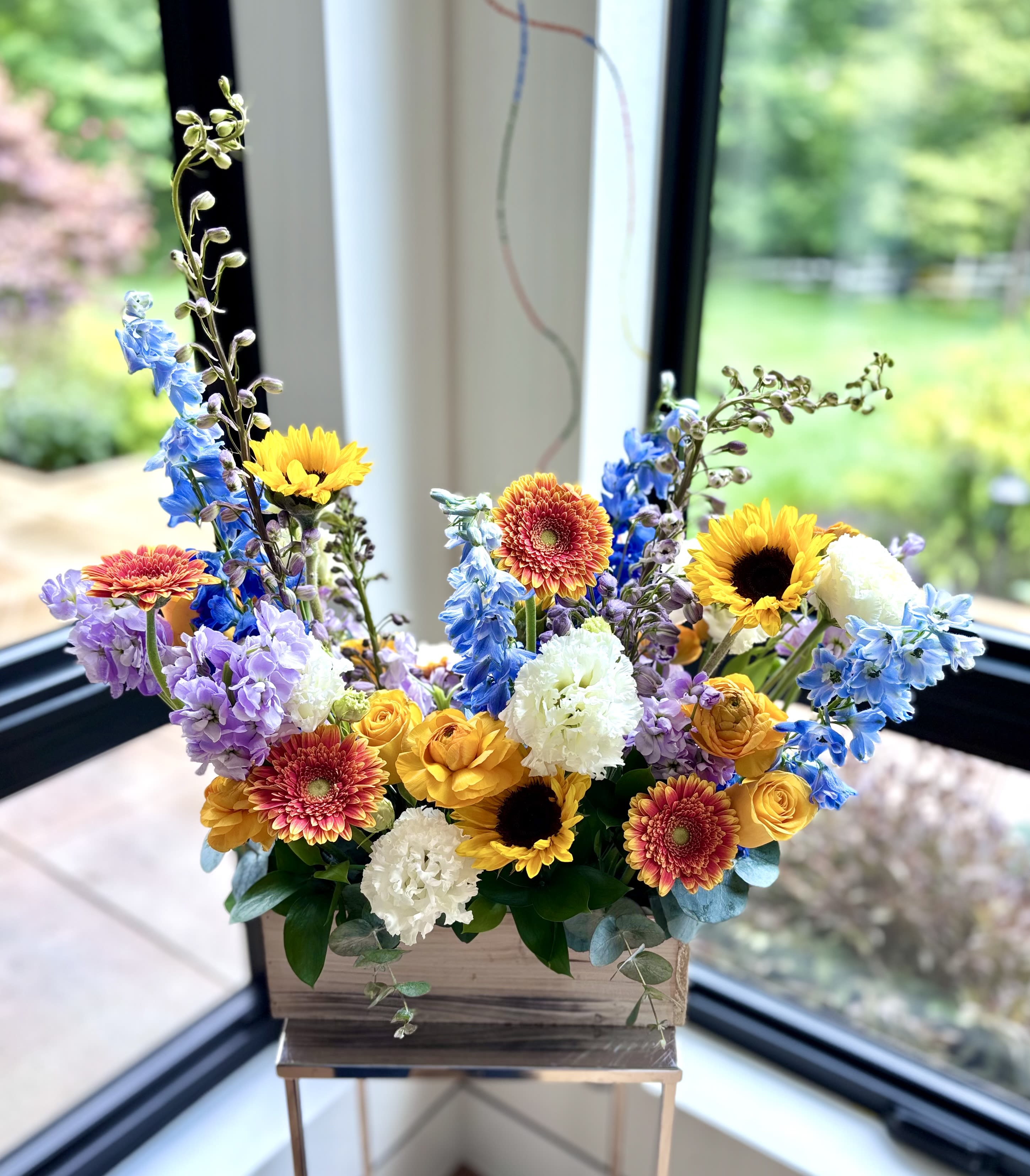 Sunny Bliss - A flower gift box full of sunshine ... with sunflowers, gerberas, yellow roses and yellow ranunculus complemented with lavender stock and blue delphinium in a 12inch long wooden planter. Invite this flower sunshine into your home.