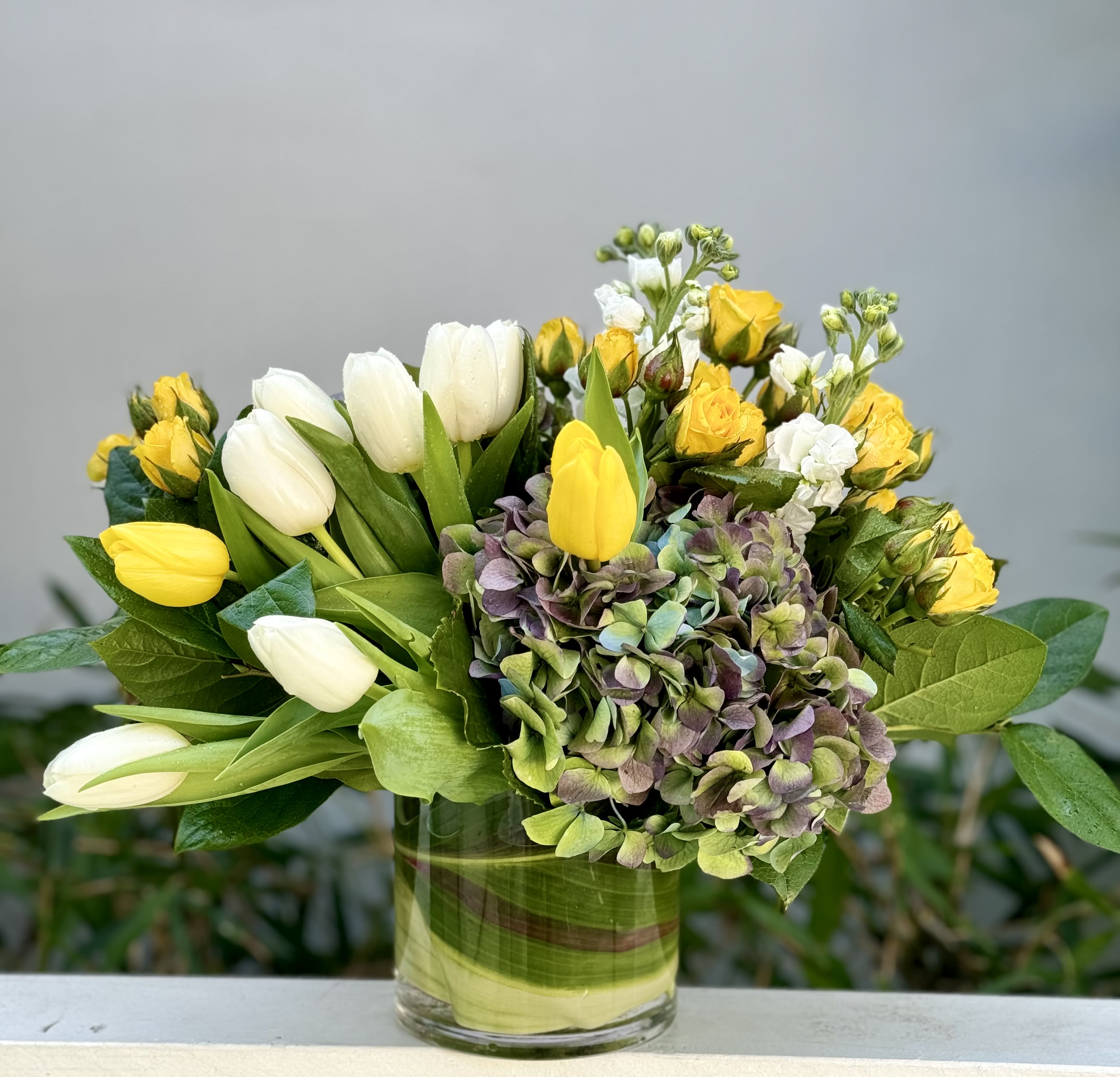Low glass vase of yellow and white tulips, yellow spray roses, stock, and hydrangea blooms.