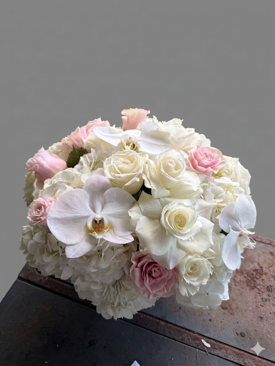 Like a virgin - Ivory and blush roses mingled with white phalaenopsis orchids and cloud-like hydrangeas create an elegant, romantic bridal bouquet perfect for a wedding or anniversary. This rounded, densely packed arrangement reads soft and refined-the kind of bouquet that makes the person you're celebrating feel seen and cherished on a milestone day.  Creamy white roses and pale pink roses form the core, accented by delicate white Phalaenopsis orchids and clusters of white hydrangea for fullness and gentle texture. The compact, dome-like shape is hand-tied for a polished finish and easy carrying; the palette of white, pink, and mixed soft tones keeps the look timeless and photo-ready.  Ideal for brides, vow renewals, or someone you want to honor with classic romance, this arrangement communicates purity and tenderness without being fussy. Sizes are kept intentionally compact for a refined silhouette; care is simple-keep stems recut and water refreshed to preserve bloom life for the celebration.