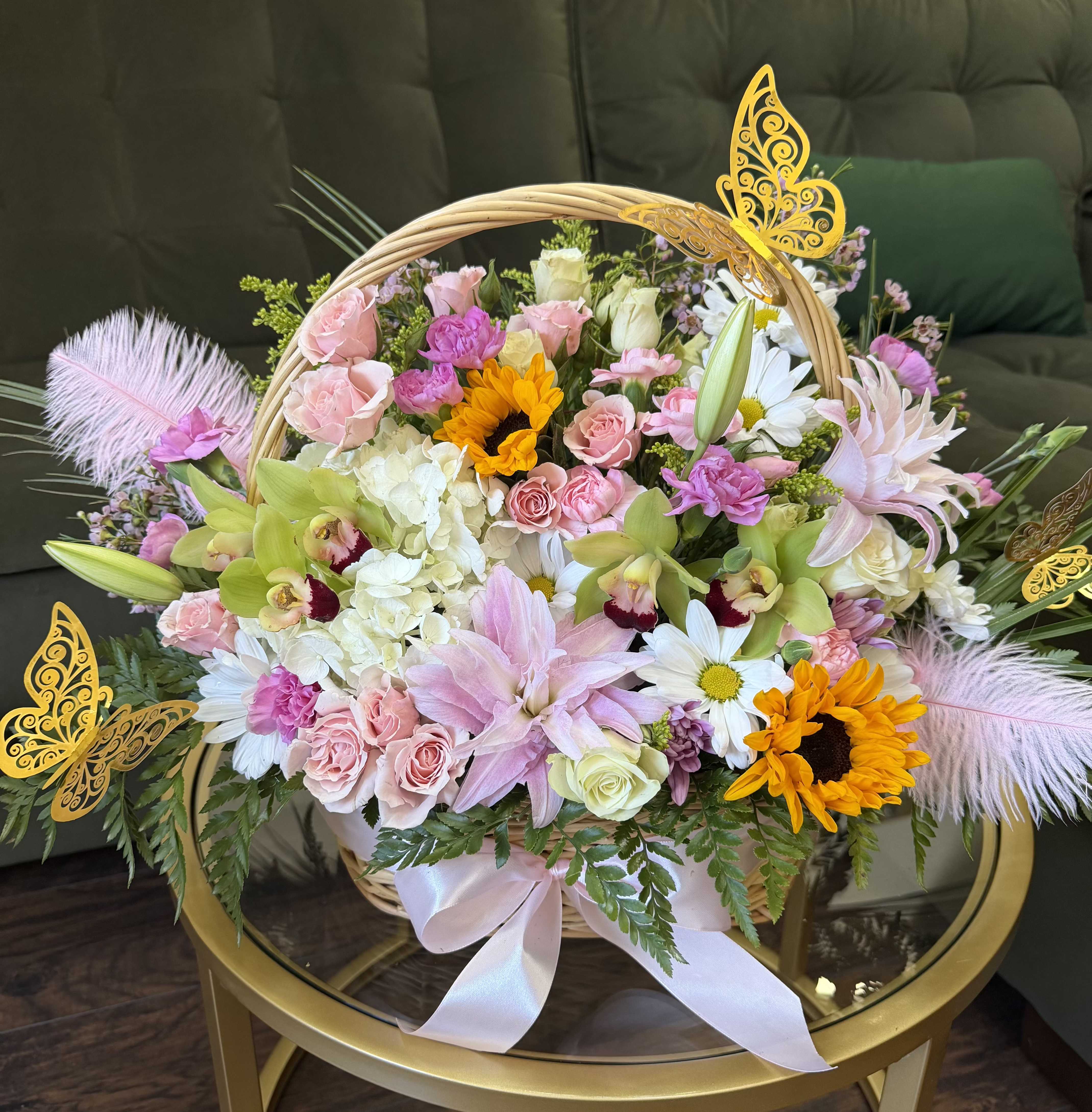 Signature Garden Basket - An enchanting garden-style luxury basket overflowing with delicate blush and pastel blooms, designed to feel whimsical yet refined. This romantic composition features soft pink roses, graceful lilies, creamy hydrangea, elegant orchids, cheerful sunflowers, and dainty white daisies, artfully layered with lush greenery. Finished with a silky blush ribbon and adorned with golden butterfly accents and pink feathers, this arrangement evokes lightness, joy, and timeless romance.  Perfect for celebrating love, welcoming new beginnings, or making a truly unforgettable gesture. Each basket is handcrafted with thoughtful detail and luxurious texture, ensuring a one-of-a-kind floral experience.