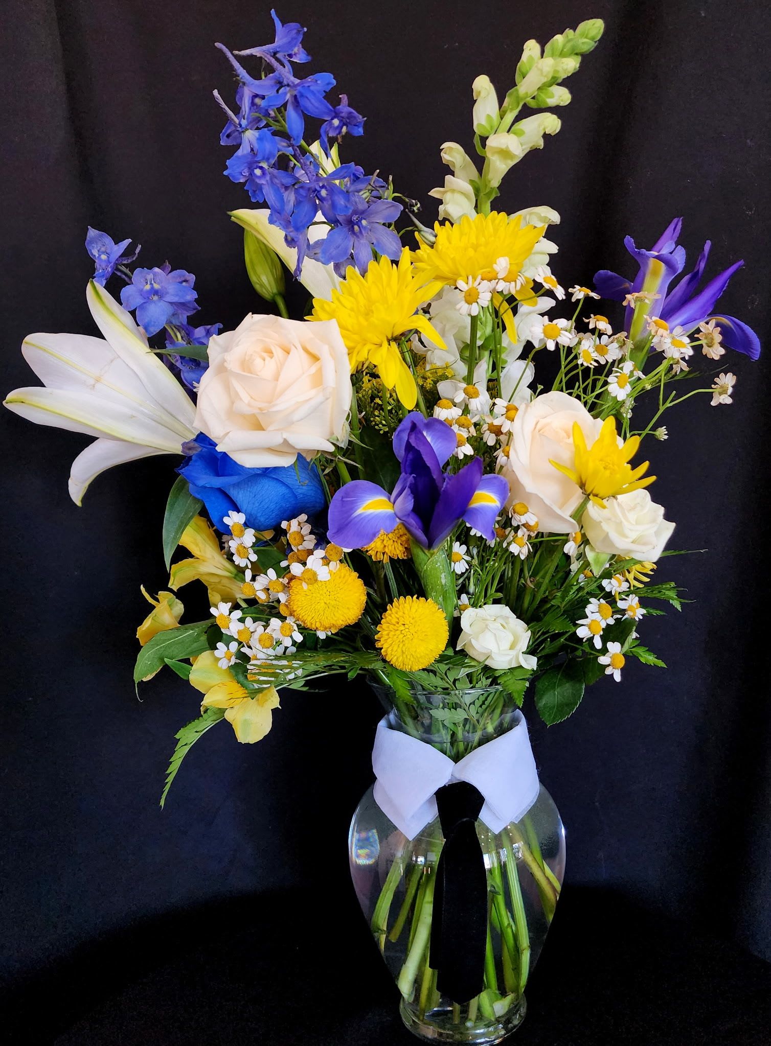 For the Handsome Father - Give to your handsome father/husband or friend this beautiful arrangement to celebrate this special day. This arrangement includes white roses, a single blue rose, irises, snapdragon, white Lillies, mums, alstroemeria, delphinium, chamomile, solidago and leather greens. It also includes the collar and tie as seen in the picture for added style.  The glass vase is 8" tall and the arrangement is 21" Height x 13" Width.