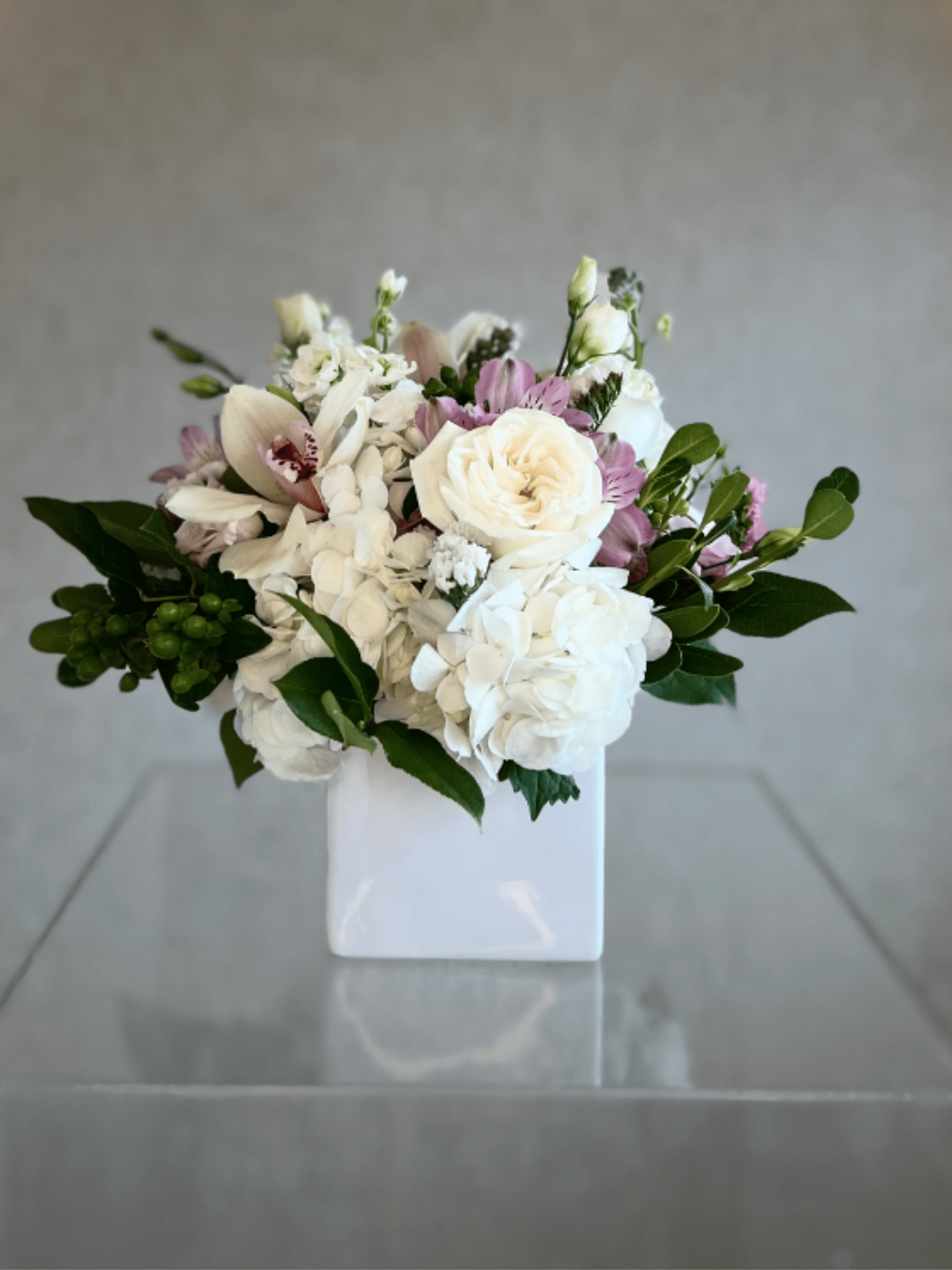 Cymbidium Daydream - A fresh, romantic tabletop arrangement that reads soft and refined-perfect as a wedding centerpiece or an elegant sympathy arrangement. This flower arrangement pairs white hydrangeas and white roses with pale pink alstroemeria and lisianthus to create a compact, rounded bouquet that feels both intimate and lifted. The white ceramic vase keeps the look modern and understated while the overall palette-white, pink, and green-sends a calm, thoughtful message to the person you're honoring.  Flowers are layered densely for a plush, tactile surface: billowy hydrangea clusters, creamy rose petals, and delicate pink alstroemeria and lisianthus intermix with glossy dark foliage for subtle contrast. The arrangement sits low and balanced in a glossy square white vase, making it ideal for tabletops, mantels, or a reception table where a restrained yet visible floral statement is needed. Sized to be approachable and easy to place, it requires only fresh water-change it every other day and trim stems slightly on re-cut to keep blooms looking their best.