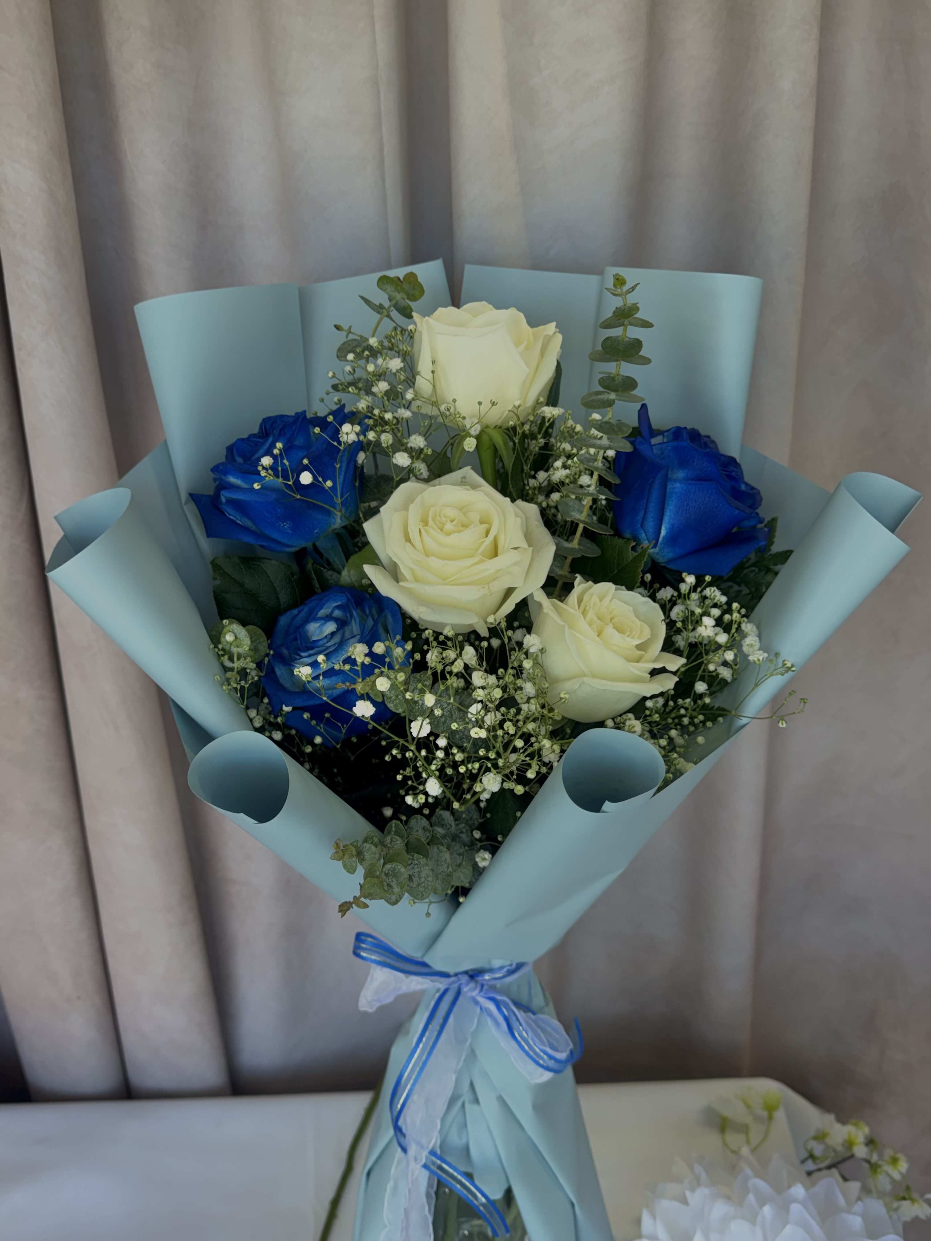 ocean blues - Vivid blue roses mingle with classic white roses in this Ocean Blues bouquet, a striking birthday bouquet or celebratory gift that reads as both romantic and refreshingly modern. The cool-toned pairing of blue and white is especially fitting for graduations, baby showers, or anyone who loves a serene coastal palette-she'll feel noticed and celebrated the moment the wrapping is opened.  Arranged in a rounded, hand-tied shape, the bouquet includes several white roses, vivid blue roses, delicate white filler flowers, and eucalyptus-like greenery for soft texture and movement. Light blue wrapping paper and coordinating blue and white ribbons finish the look, keeping the presentation polished and easy to carry. The mix of crisp whites, deep blues, and soft greens creates a clean, sculpted silhouette that stands out without being fussy.  This is the kind of bouquet that reads as thoughtfully chosen-the cool colors convey calm and admiration for graduations, anniversaries, or simply saying "I'm thinking of you." Hand-delivered and neatly wrapped, it arrives ready to display; keep blooms in fresh water and trim stems for longest enjoyment.
