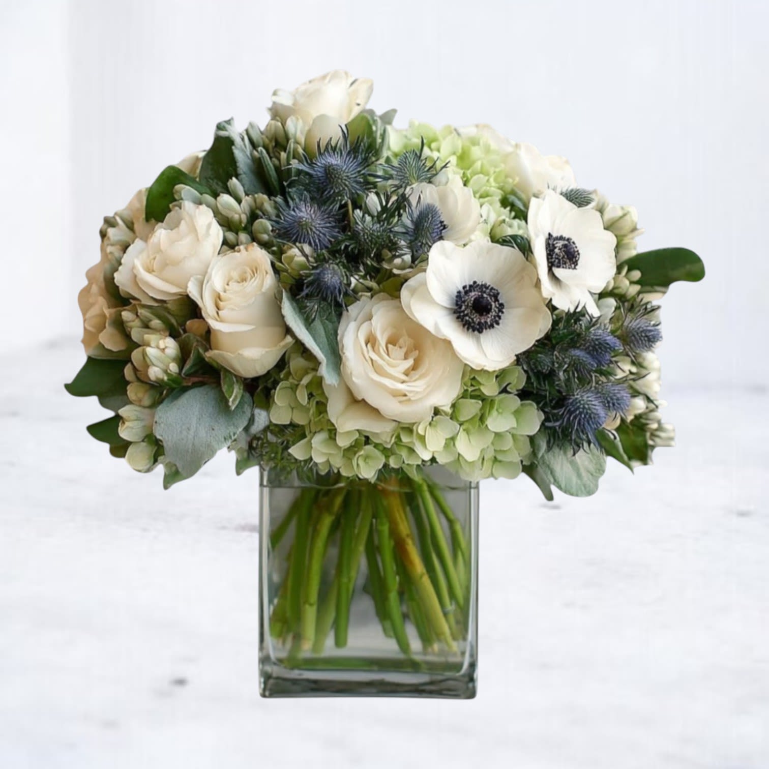 Flourish &amp; Frond - Elegant yet effortlessly natural, Flourish &amp; Frond blends creamy roses, crisp white anemones, fresh green hydrangea, and textured blue thistle in a beautifully balanced composition. Layers of eucalyptus and curated greenery add depth and movement, creating a design that feels polished but never overdone. Arranged in a clear glass vase, this low, lush composition offers a gentle presence without overwhelming the space. Its neutral palette and organic movement make it a thoughtful choice for sending sympathy flowers to the home, honoring a life well lived, or simply letting someone know they are not alone.  Whiskey Wallflower is a design-forward florist serving Magnolia, The Woodlands, Montgomery, Willis, Conroe, Spring, Tomball and surrounding Houston-area communities. We specialize in thoughtfully composed, modern floral arrangements with a focus on premium blooms, intentional design, and reliable same-day delivery. Every piece is created with care to make life’s moments feel elevated, personal, and unforgettable.