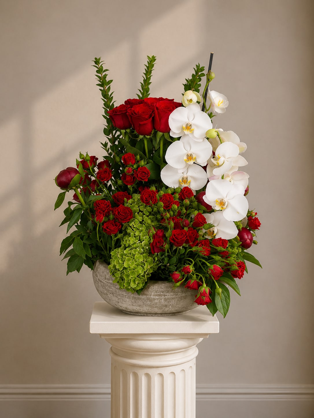 Exquisite Embrace - Deep clusters of deep red roses meet cascading white Phalaenopsis orchids in a dramatic anniversary bouquet built to make someone feel deeply cherished. This flower arrangement for anniversaries, romantic occasions, or refined everyday gifting uses strong color contrast-saturated red and crisp white-to create an immediate emotional impression: romantic, polished, and unmistakably special.  The design pairs lush red roses with bright green hydrangea-style blooms and upright greenery for texture and height, forming a layered, sculptural silhouette. White Phalaenopsis orchids trail gracefully for a soft, elegant contrast, and the whole composition is presented in a rounded gray ceramic bowl vase that grounds the arrangement with a modern, upscale feel. The asymmetrical shape and varied heights give it a statement-making presence ideal for a dining table, entryway, or bedside display.  Give this to the person you're thinking of when you want them to feel celebrated, adored, or seen-especially for anniversaries or moments of romance. The sturdy ceramic bowl keeps stems secure; to extend bloom life, refresh the water as needed and keep the arrangement out of direct heat and drafts.