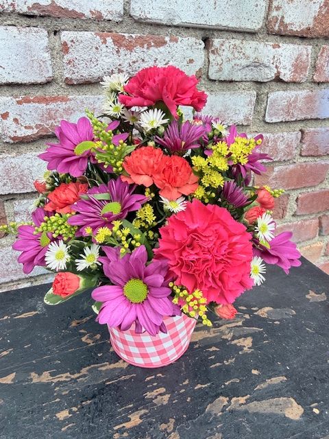 Petal Picnic - Bright, playful color pops make this Petal Picnic flower arrangement an instant mood lifter - perfect as a birthday bouquet, a cheerful Mother's Day surprise, or a just‑because pick-me-up. The combination of vivid pink carnations and large purple daisy-like blooms reads joyful and sincere, while small white daisies and clusters of sunny yellow filler flowers give the mix a fresh, springtime brightness that also works for thank-you or I'm‑sorry gestures.  Packed into a rounded, compact design, the stems sit snugly in a charming pink gingham container that gives the piece a casual, giftable look - ideal for summer barbecues, a thoughtful Women's Day bouquet, or a Parent's Day surprise. Textures range from the ruffled carnations to the crisp daisy petals, creating lively contrast and visual depth that makes the recipient feel truly seen.  This arrangement arrives ready to display; its compact size fits on tabletops, desks, or kitchen counters so she'll enjoy the color right away. Order for same‑day delivery when available to celebrate birthdays, spring moments, or those spontaneous "thinking of you" occasions that deserve something bright and personal.