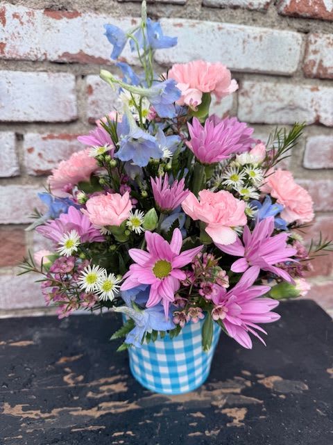 Blooming Blue Happiness - Soft, cheerful pastels make this Blooming Blue Happiness flower arrangement a bright everyday bouquet perfect for birthdays, spring celebrations, or a heartfelt thank-you. The casual, homey look feels like a friendly visit in floral form-ideal for the person you want to make smile and feel genuinely seen.  This compact mixed bouquet features ruffled pink carnations, lavender and purple daisy-like blooms, pale blue flowers, and tiny white filler blossoms for an airy, garden-fresh texture. Stems are gathered in a blue-and-white gingham metal container that adds country charm and keeps the design neat and display-ready on a kitchen table or desk.  Thoughtful but unfussy, this arrangement is the kind of gift that reads as both celebratory and comforting-great for a neighbor, coworker, or friend. The ceramic gingham container arrives ready to display; refresh water daily and trim stems slightly if re-arranging to keep blooms looking their best.