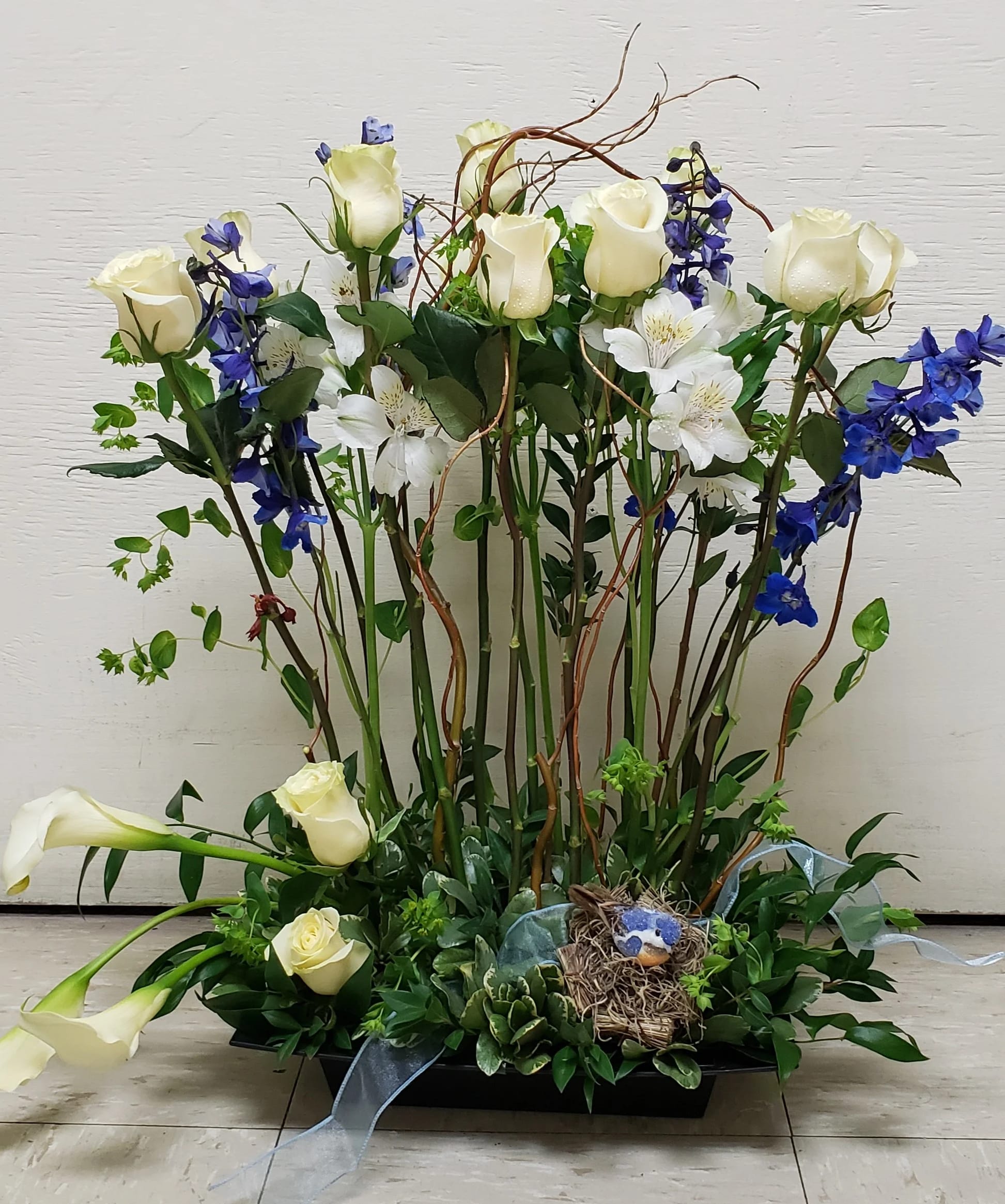 Tall arrangement of white roses, calla lilies, and blue flowers with a small bird nest in a low tray