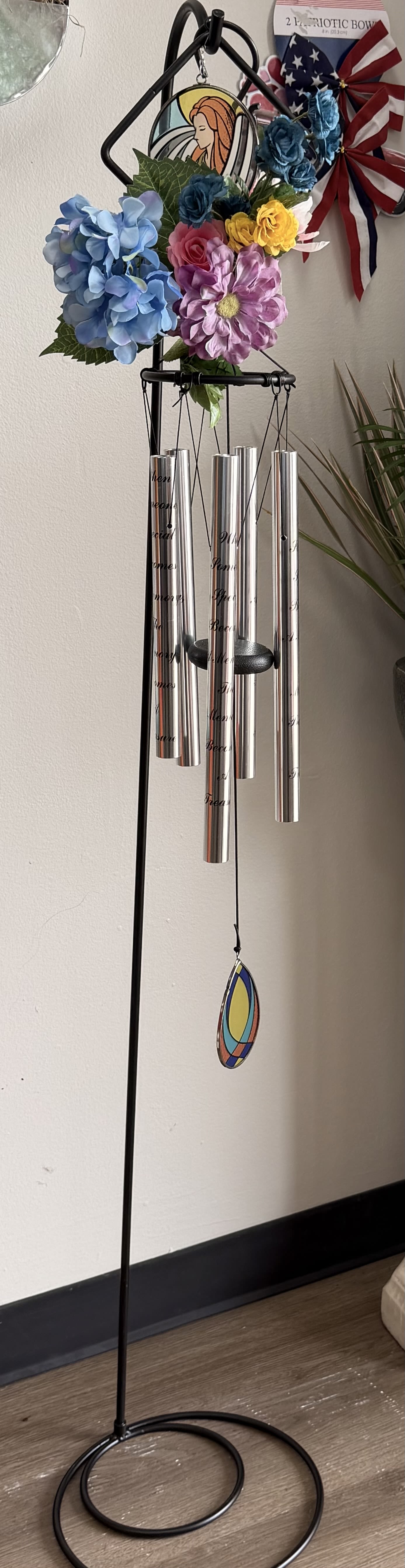 * Faith &amp; Remembrance Wind Chime - A bright floral topper crowns a polished silver wind chime for a memorial or sympathy gift that feels hopeful rather than heavy - a faith and remembrance wind chime designed to comfort and celebrate a life. As a sympathy arrangement and outdoor memorial accent, this flower arrangement pairs the calming sound of metal chimes with cheerful blue, pink, purple, and yellow blooms to create a peaceful focal point for a garden, porch, or remembrance corner.  The piece features lifelike hydrangeas, roses, daisies and complementary filler blooms arranged on a decorative hanging cluster above the chimes. A tall black metal stand displays the hanging set of polished silver wind chimes and a small stained-glass-style ornament beneath, while a patriotic ribbon bow near the top adds a thoughtful touch. The colors read bright and uplifting, and the mixed textures of rounded hydrangea clusters, soft rose petals, and daisy centers give visual depth even from a distance.  Ideal for honoring a loved one's memory, marking a graveside visit, or offering sympathy to someone who prefers an enduring keepsake, this arrangement comes ready to display in a yard or on a covered porch - no watering required. It's the kind of gift that lets the person you're thinking of hear gentle reminders of your care whenever the breeze moves the chimes.