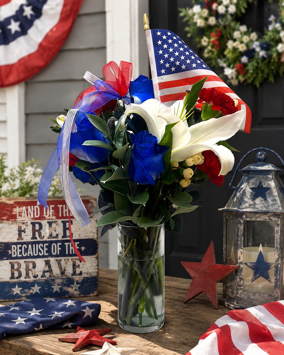 Liberty Bouquet (Limited Time Only) - A bold, patriotic display built to honor and celebrate-this flower arrangement is a Fourth of July and Memorial Day bouquet that feels both ceremonial and heartfelt. The Liberty Bouquet (Limited Time Only) pairs white lilies with blue roses and red carnations so the person you're thinking of will feel seen and celebrated with classic American colors and a festive, upright silhouette.  Arranged in a clear glass vase, the mixed-color stems form a full, clustered presentation accented by red, white, and blue decorative ribbon and a small American flag tucked into the stems. The composition reads traditional and substantial: fragrant white lilies provide height and scent, blue roses add an unexpected cool tone, and red carnations give durable, vivid body. The product image also shows complementary Americana hard goods-a lantern and rustic wooden sign-suggesting the arrangement works beautifully alongside patriotic décor.  Perfect for honoring a veteran, decorating a holiday table, or sending a meaningful message on Memorial Day or Independence Day, this bouquet makes a proud, ceremonial gift. Vase-ready and made to display, follow the included Care and handling QR code for watering and longevity tips to keep the flowers looking their best through the celebrations.