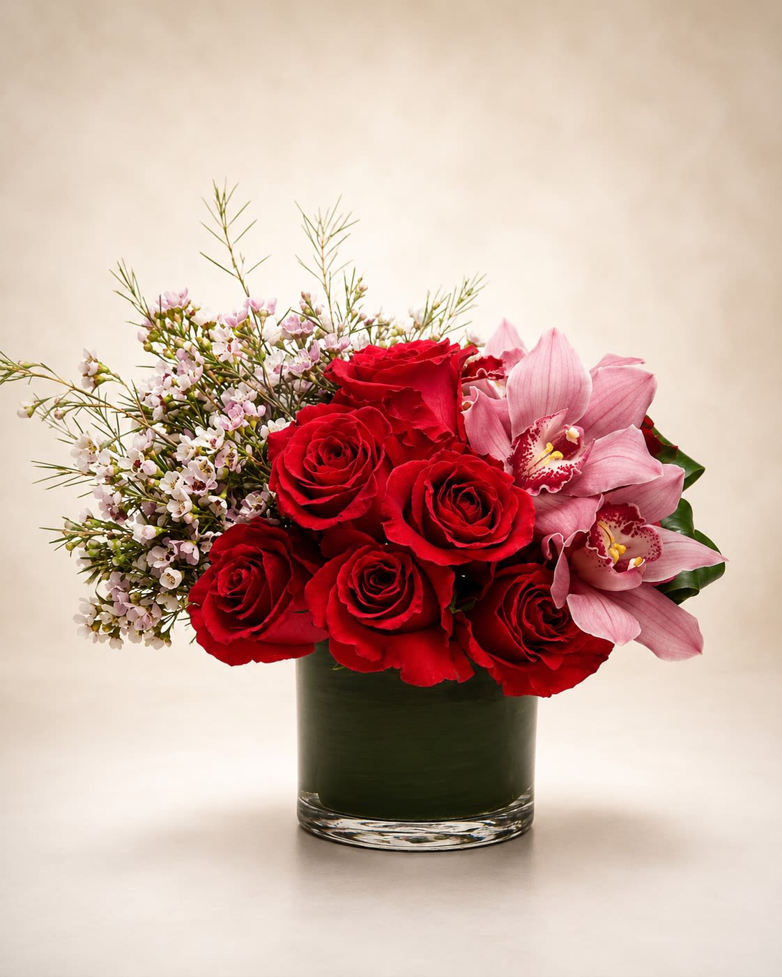 Velvet Orchid Romance - A romantic luxury floral arrangement featuring premium red roses paired with soft pink cymbidium orchids and delicate seasonal waxflower designed in a modern leaf-lined cylinder vase. Velvet Orchid Romance blends timeless romance with graceful elegance, combining rich crimson blooms and soft blush orchid accents for a sophisticated garden-inspired presentation.  This arrangement is designed with lush premium roses as the focal point, complemented by exotic orchids and airy seasonal textures that create movement and softness throughout the design. The contrast of deep red florals against delicate blush tones gives the arrangement a refined upscale aesthetic perfect for anniversaries, romantic gestures, birthdays, luxury gifting, or elegant celebrations.  Due to seasonal and market availability, certain orchid varieties, accent blooms, or greenery may occasionally vary while maintaining the arrangement’s romantic color palette, fullness, and luxurious overall style. Vase style and foliage lining may also vary slightly depending on availability while preserving the same polished presentation.