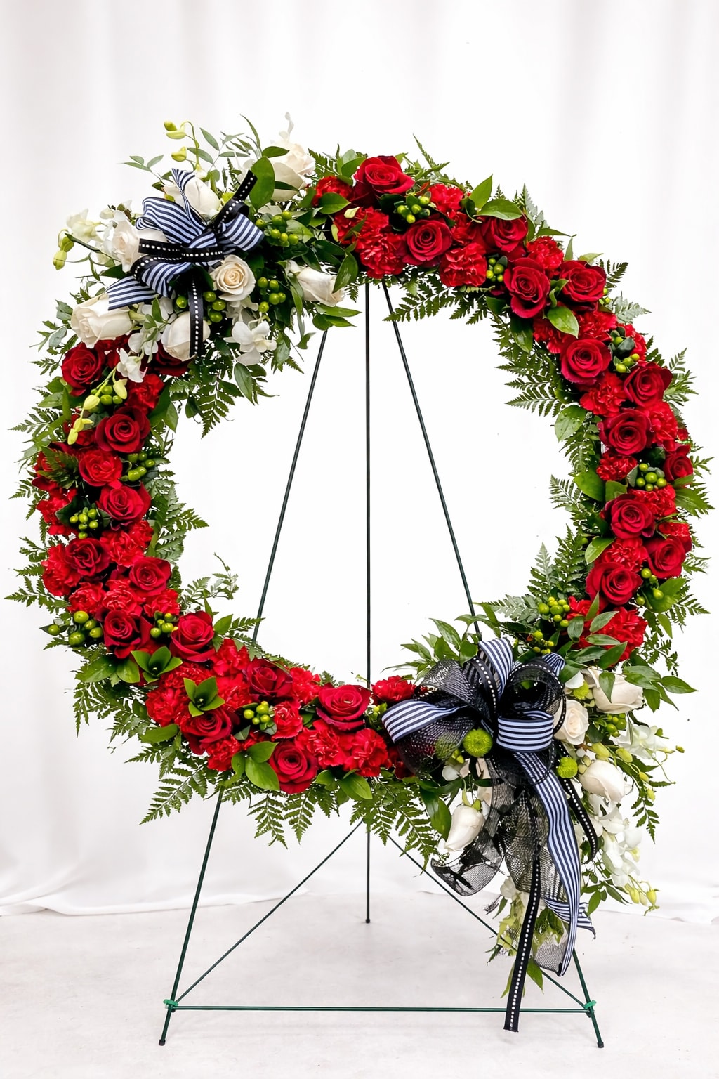 Circle of Love Tribute - A dramatic circular wreath with dense red roses and carnations makes a bold memorial tribute-perfect as a sympathy arrangement for a funeral or graveside service. The vivid red blooms contrasted against clusters of white roses and white filler flowers create a striking, ceremonial look that honors a loved one with dignity and presence.  Mixed greenery is woven through the wreath to add texture and depth while two formal ribbon bows in black, white and navy stripes, plus black mesh ribbon, give a respectful, finished touch. The wreath is mounted on a sturdy metal easel stand for easy display at services and viewings; its size and fullness ensure it reads clearly from a distance and conveys heartfelt remembrance.  This arrangement is intended to comfort the person you're remembering and communicate respect to family and friends-an unmistakable way to say you care. Please note the wreath's substantial construction and easel mounting when selecting delivery or pickup for a funeral home or graveside service.