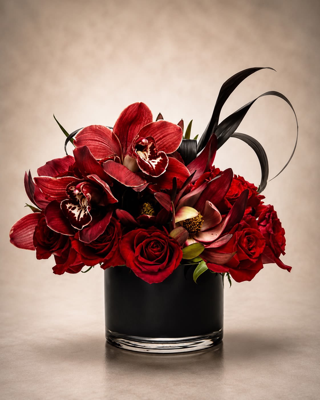 Noir Rouge Orchidée - A dramatic couture-inspired floral arrangement featuring velvety red roses, deep burgundy cymbidium orchids, rich seasonal blooms, and sculptural dark foliage designed in a sleek black glass cylinder vase. Noir Rouge Orchidée was created with a bold modern aesthetic that blends romance, mystery, and luxury into one unforgettable statement piece.  This arrangement showcases layers of premium crimson roses paired with exotic orchids in deep wine and ruby tones, accented with artistic, flowing foliage for a sophisticated, high-fashion floral presentation. The dark palette and contemporary styling make this design perfect for luxury gifting, romantic celebrations, upscale events, anniversaries, or customers looking for something daring and elegant beyond traditional florals.  Due to seasonal and market availability, orchid varieties, specialty blooms, or artistic foliage may occasionally vary while maintaining the arrangement’s rich jewel-toned palette, dramatic movement, and luxurious overall style. Vase style and decorative foliage accents may also vary slightly depending on availability, while preserving the same modern couture presentation.